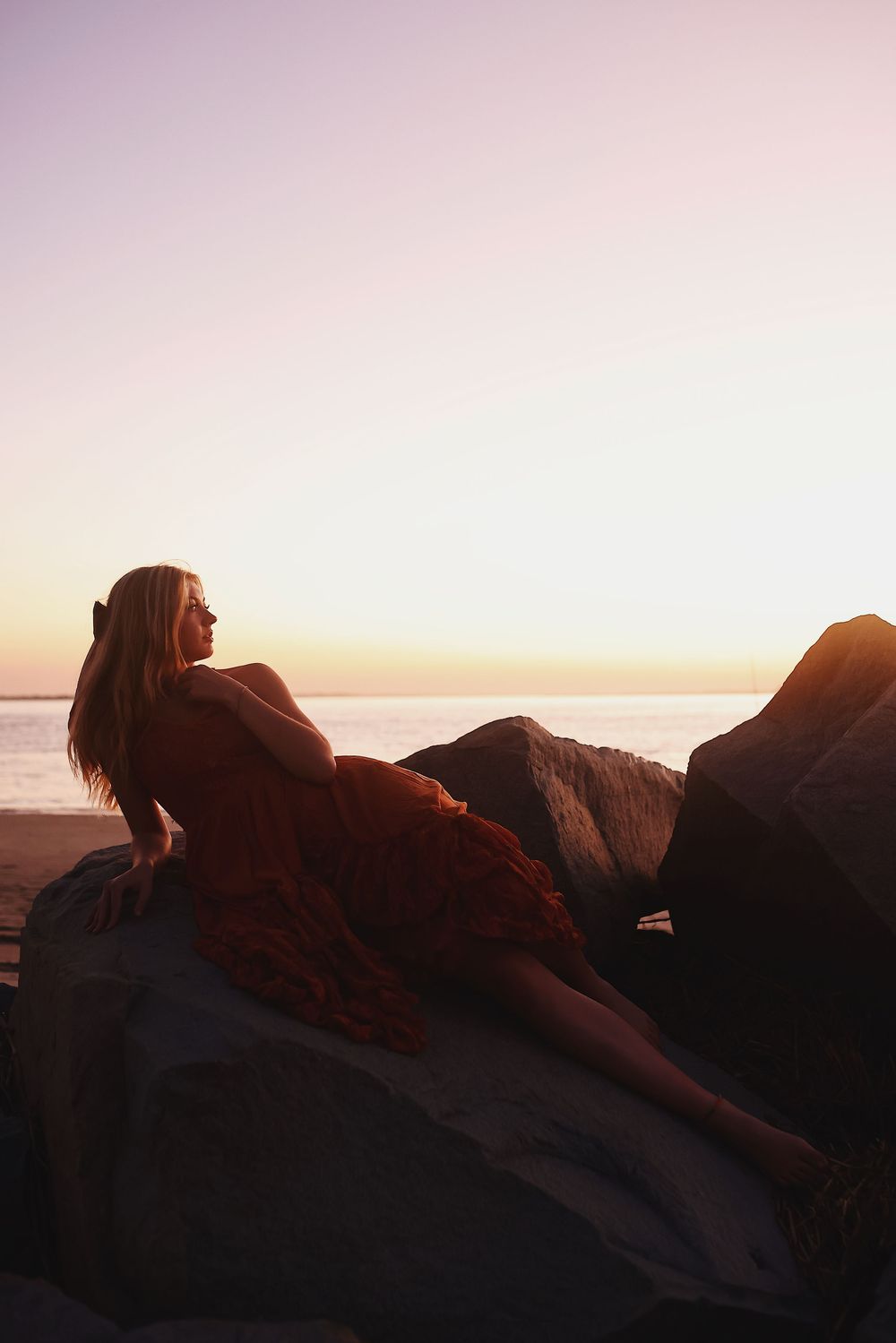 Silhouette of a person in a flowing dress reclining on coastal rocks at sunset with golden light over the ocean.