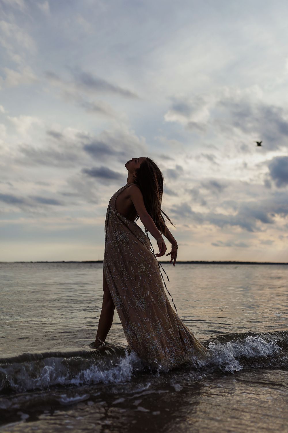 Silhouetted figure in flowing dress dances through shallow ocean waters during a moody sunset.