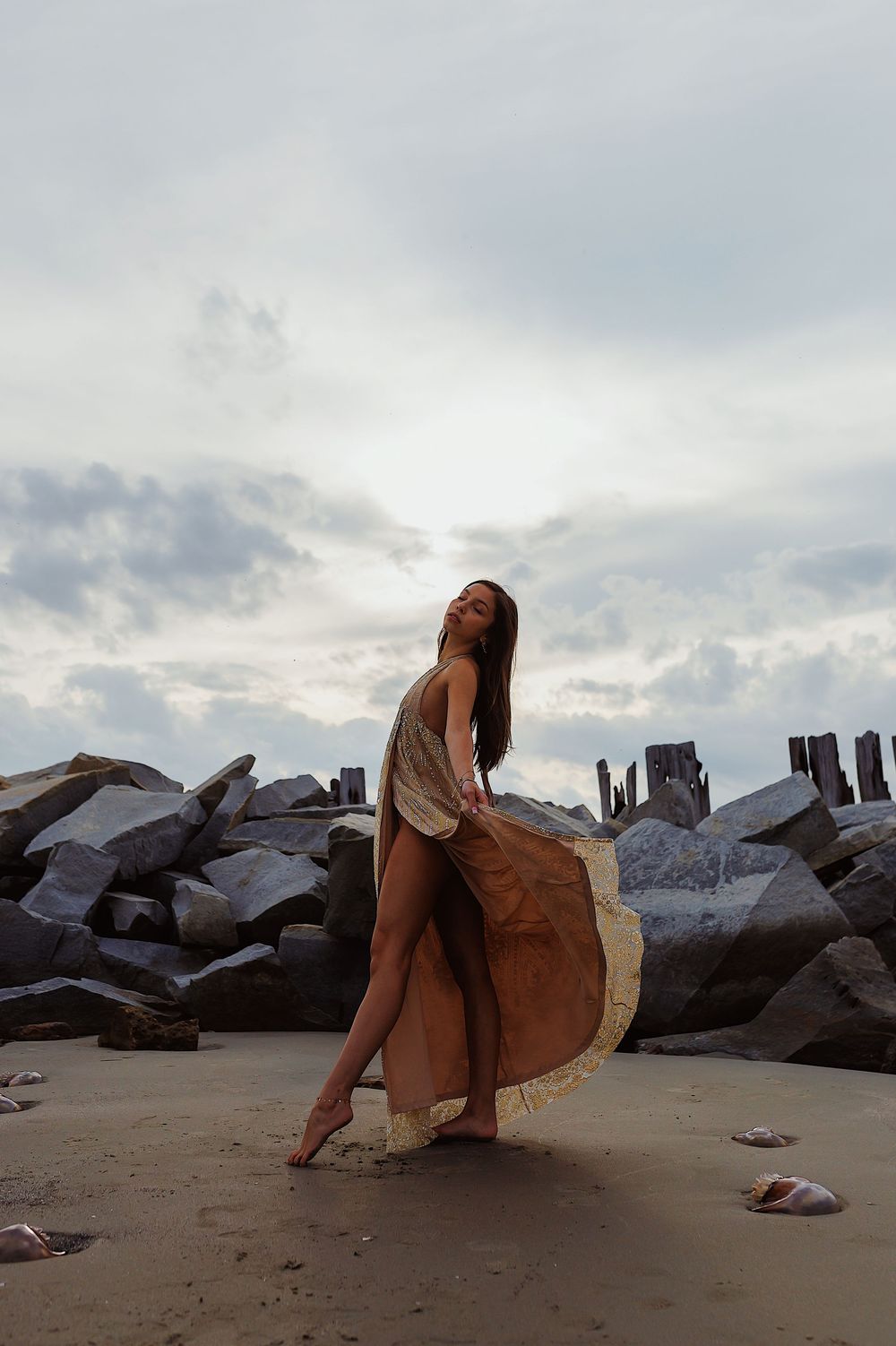 A figure in a flowing dress poses gracefully on a rocky beach at sunset with dramatic clouds in the background.