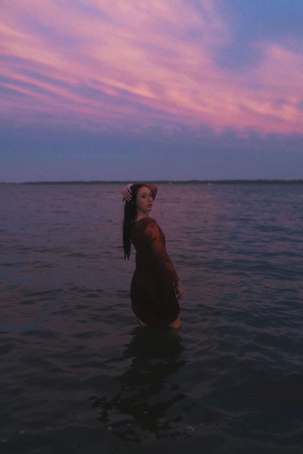 A silhouette stands in shallow water against a dramatic purple and pink sunset sky over a calm lake.