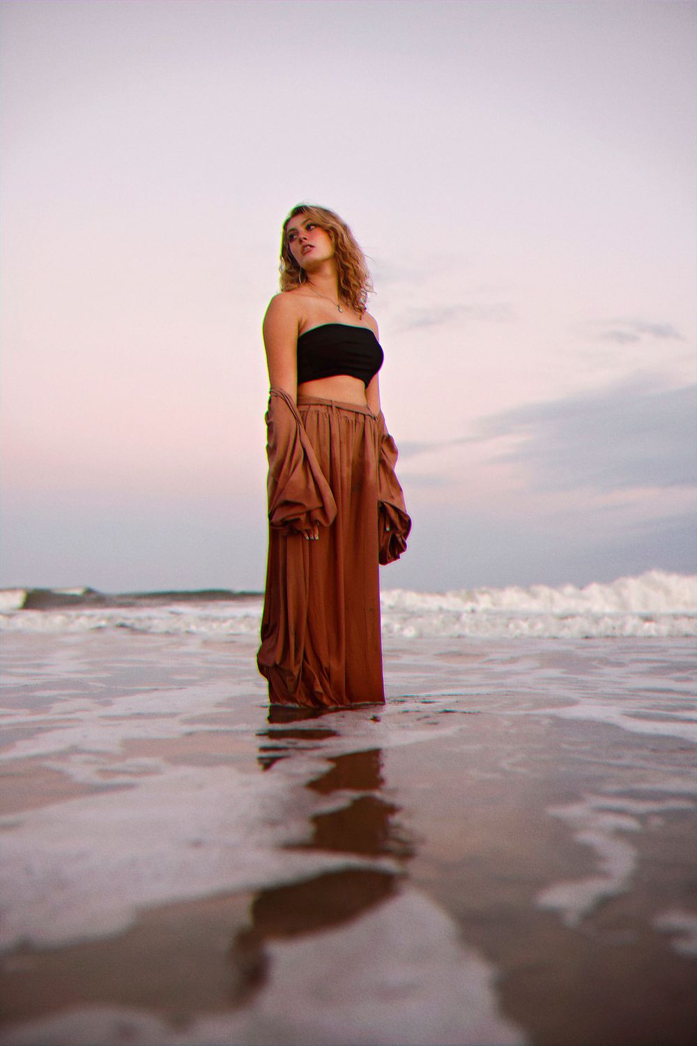 Person in black crop top and flowing brown skirt stands in shallow ocean waves during sunset.