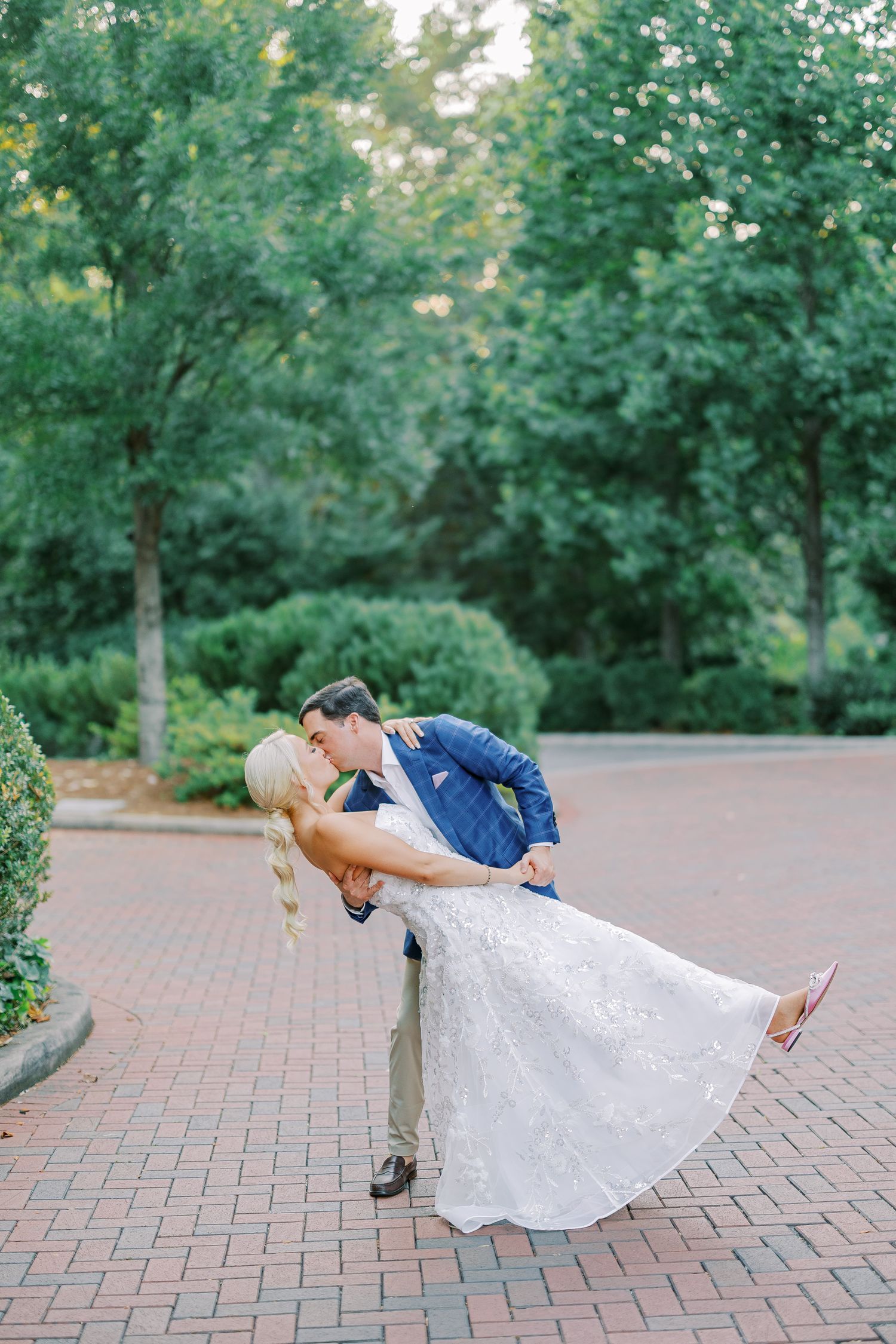 A couple in wedding attire share a romantic dip kiss on a brick pathway surrounded by lush green trees.