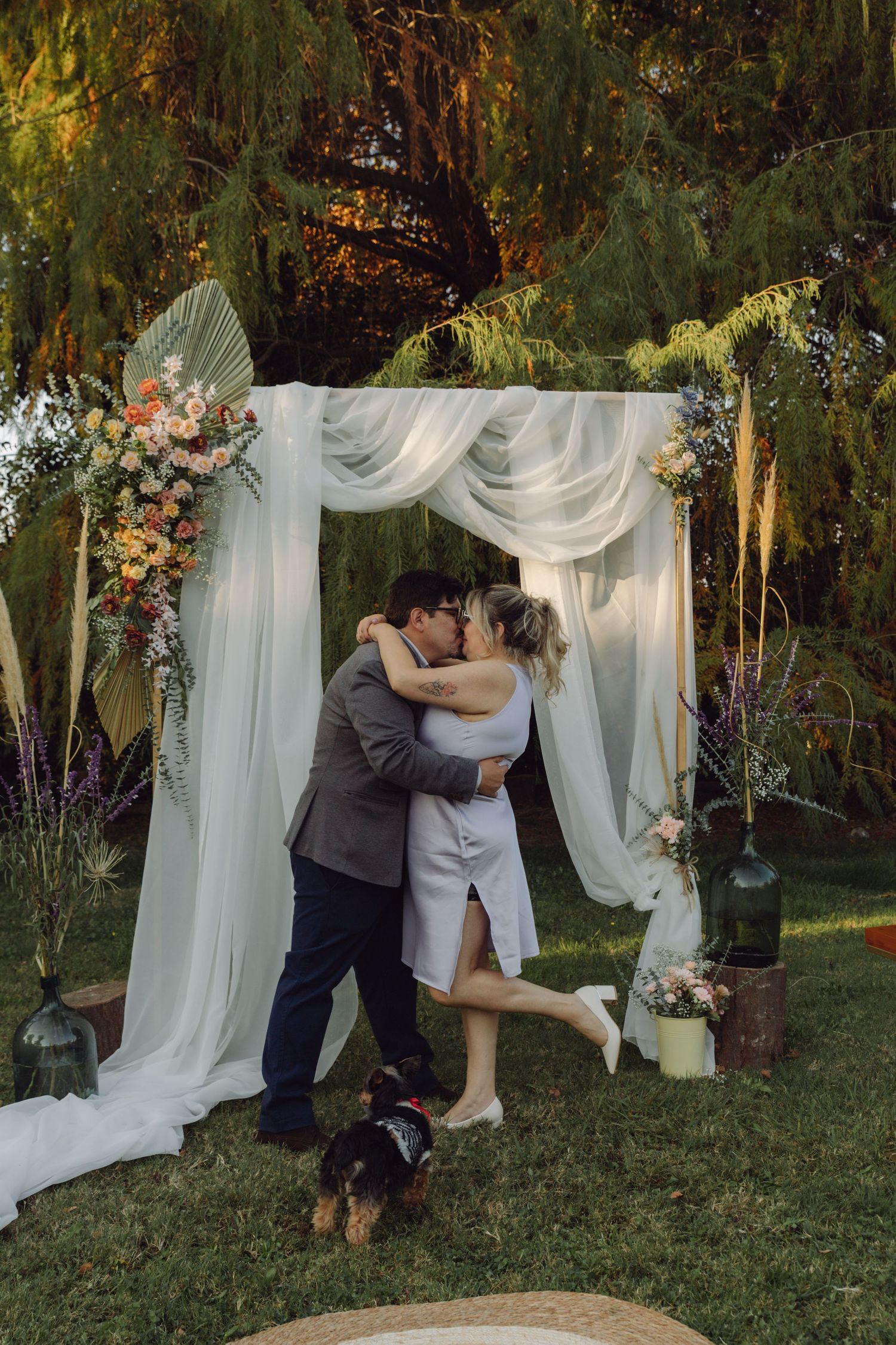 Newlyweds sharing first kiss under decorated white drape arch with flowers, small dog nearby at garden wedding.