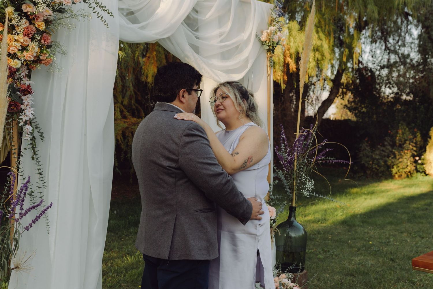 Bride and groom sharing tender moment under white draped floral wedding arch in lush garden ceremony setting.