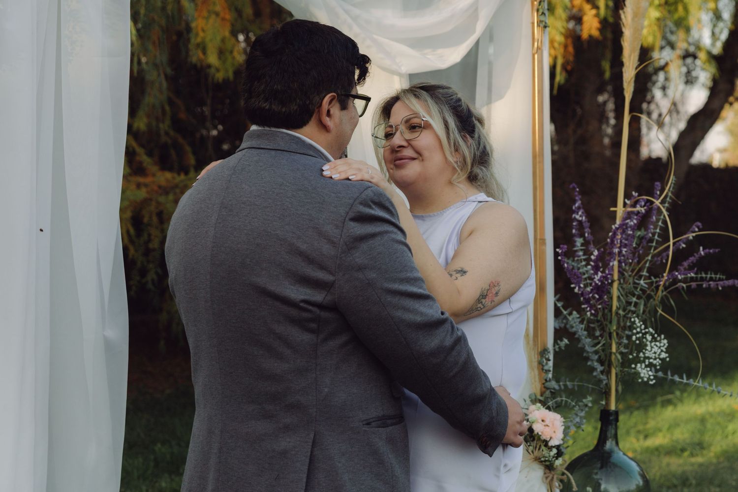 Bride smiling joyfully at groom during outdoor wedding ceremony under white draped arch with floral decorations.
