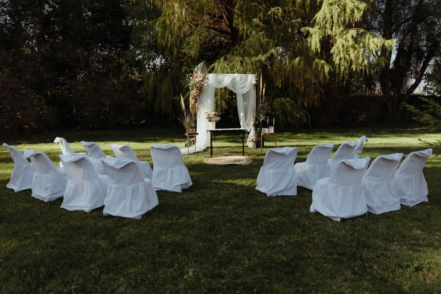 Elegant outdoor wedding ceremony setup with white chair covers and draped floral arch in a lush garden setting.