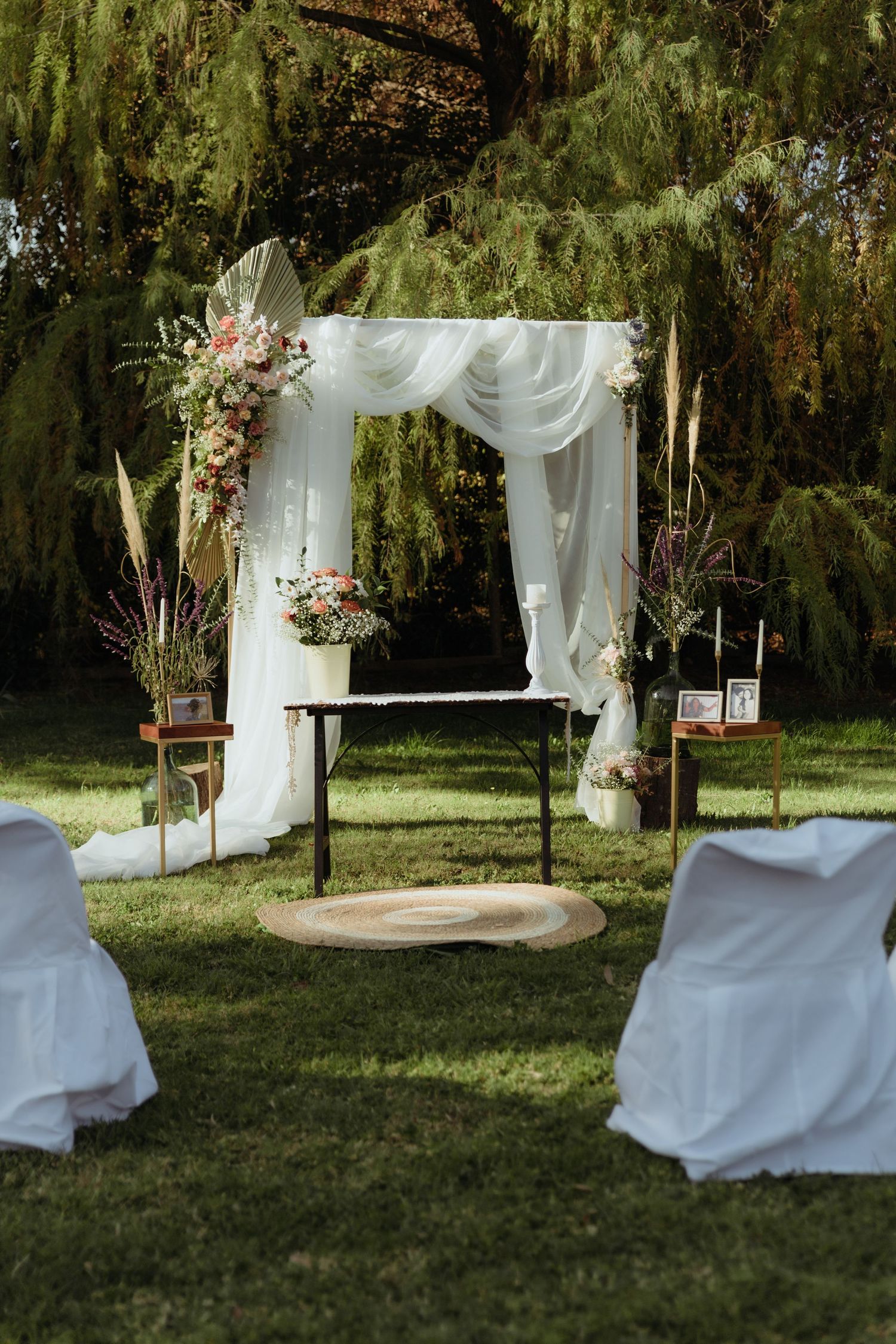 Beautiful outdoor wedding altar with white draped arch, colorful floral arrangements, and white covered guest chairs.
