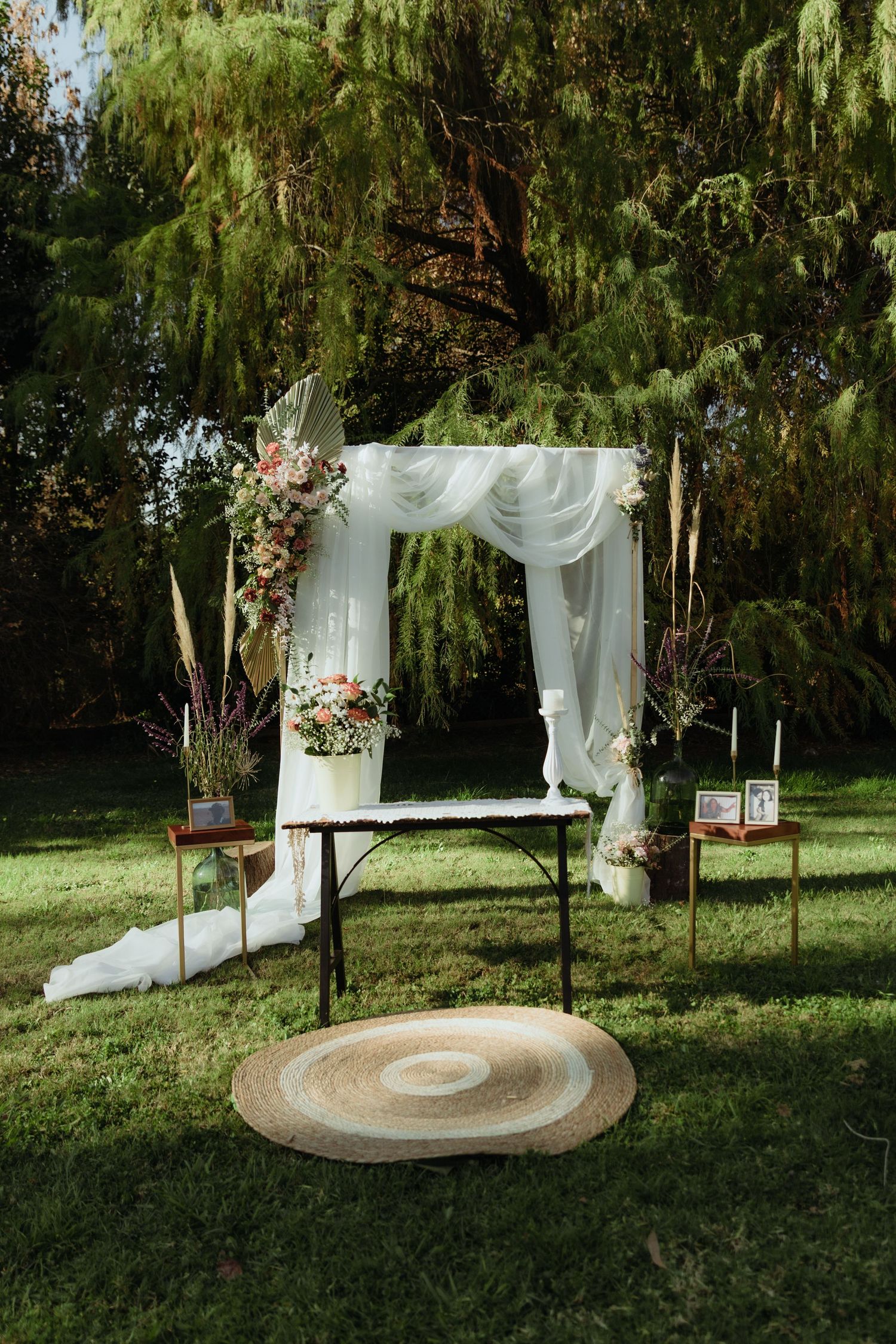 Elegant outdoor wedding ceremony arch with white draping, floral arrangements, candles, and framed photos on wooden side tables.