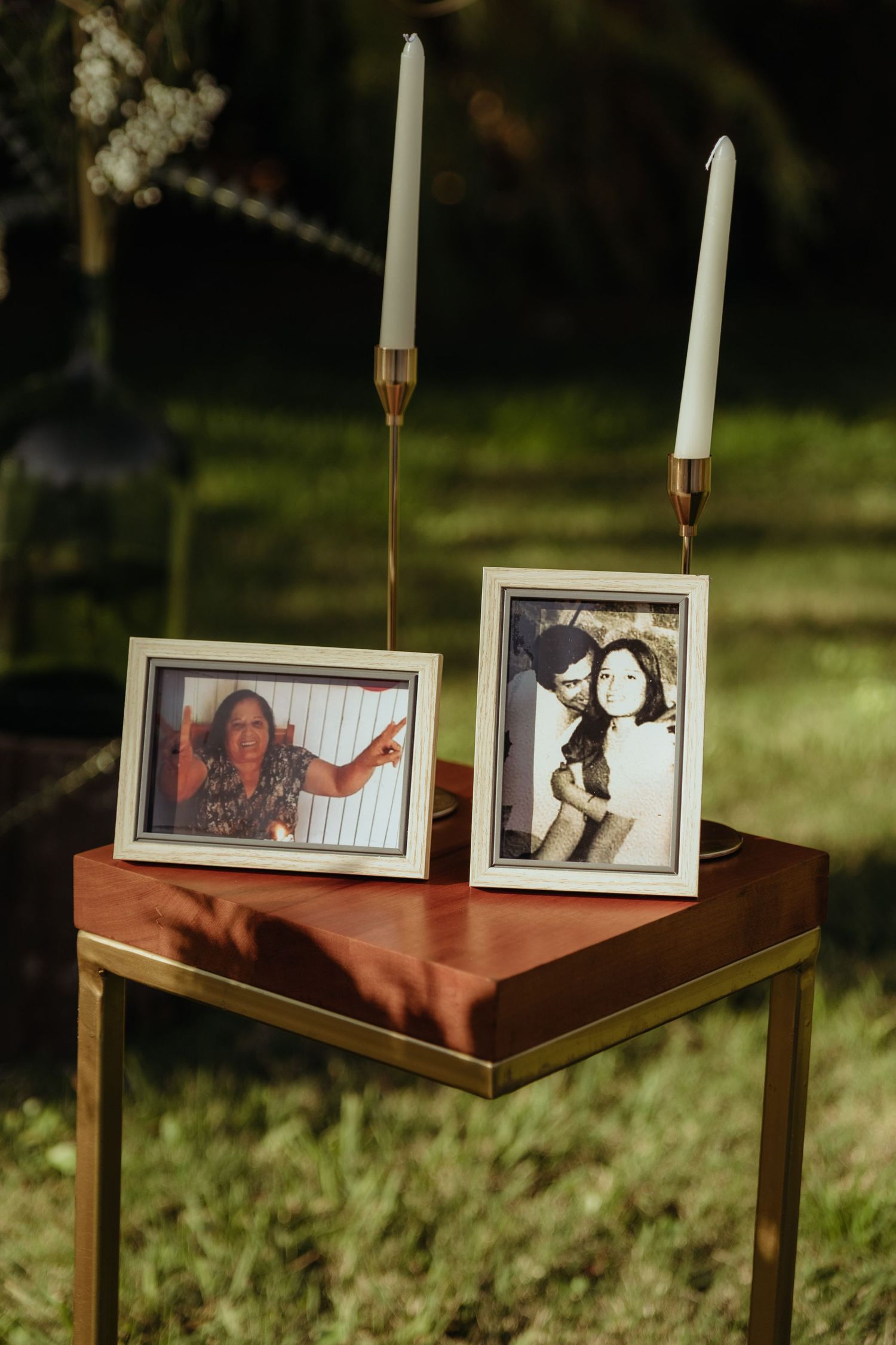 Two framed photos with white candles in gold holders displayed on a wooden table at an outdoor wedding.