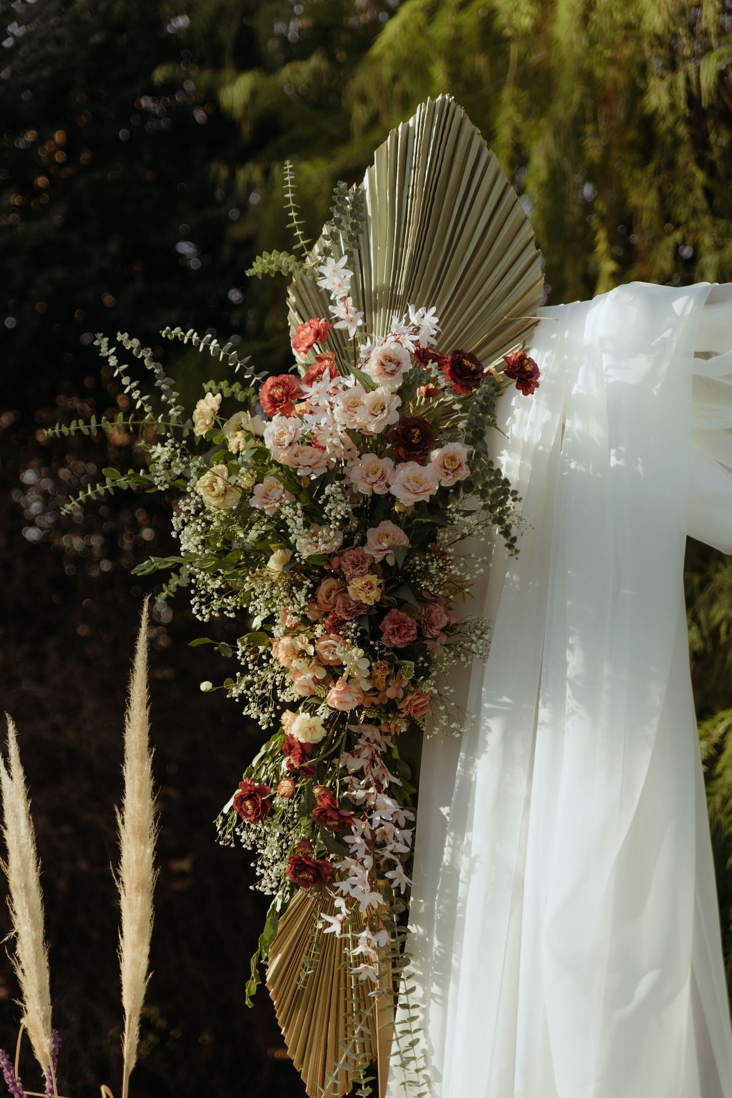 Lush boho wedding arch floral arrangement with pink, red roses, dried palm leaves, and white draping.