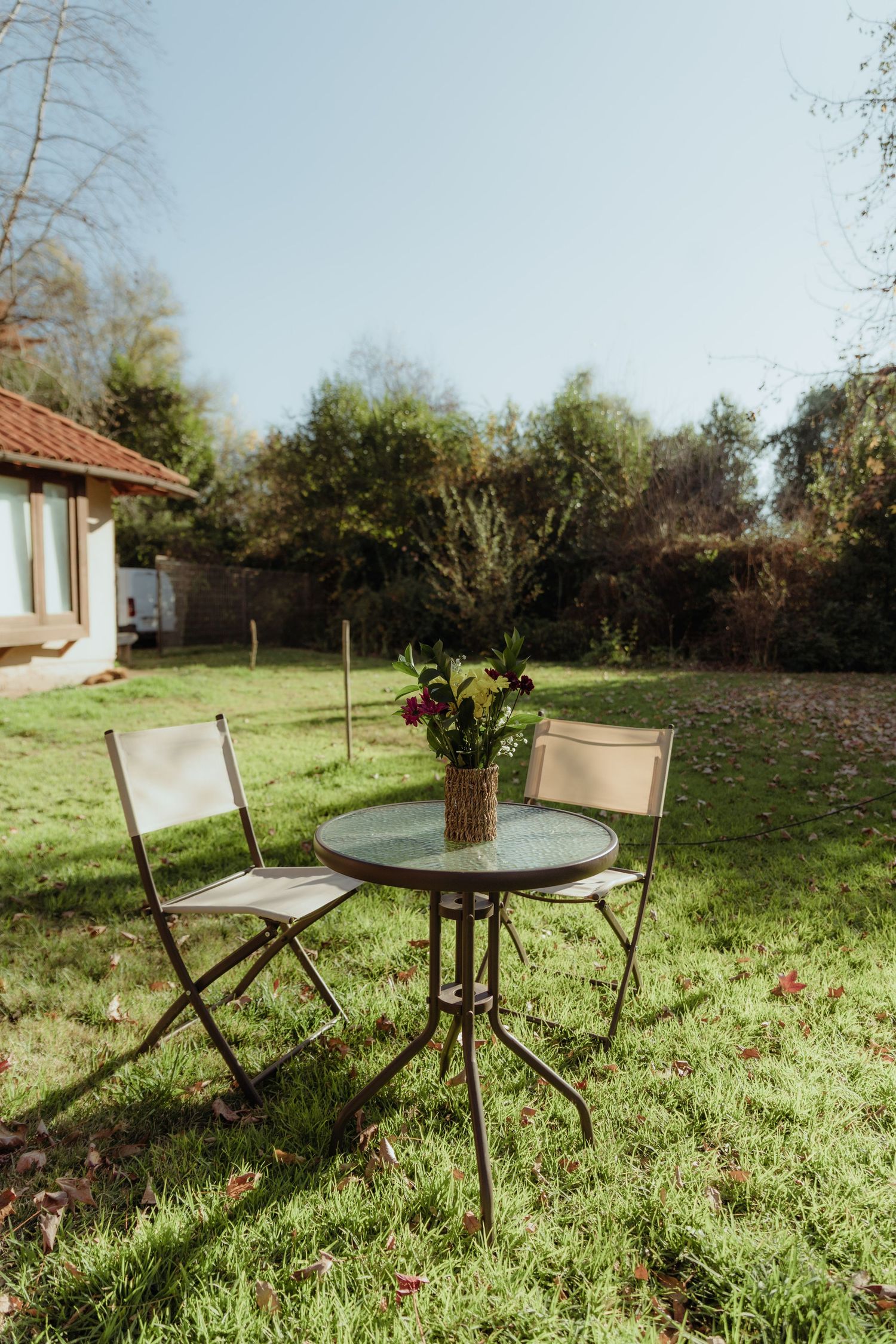 Outdoor garden seating area with two chairs and a round glass table topped with a floral centerpiece.