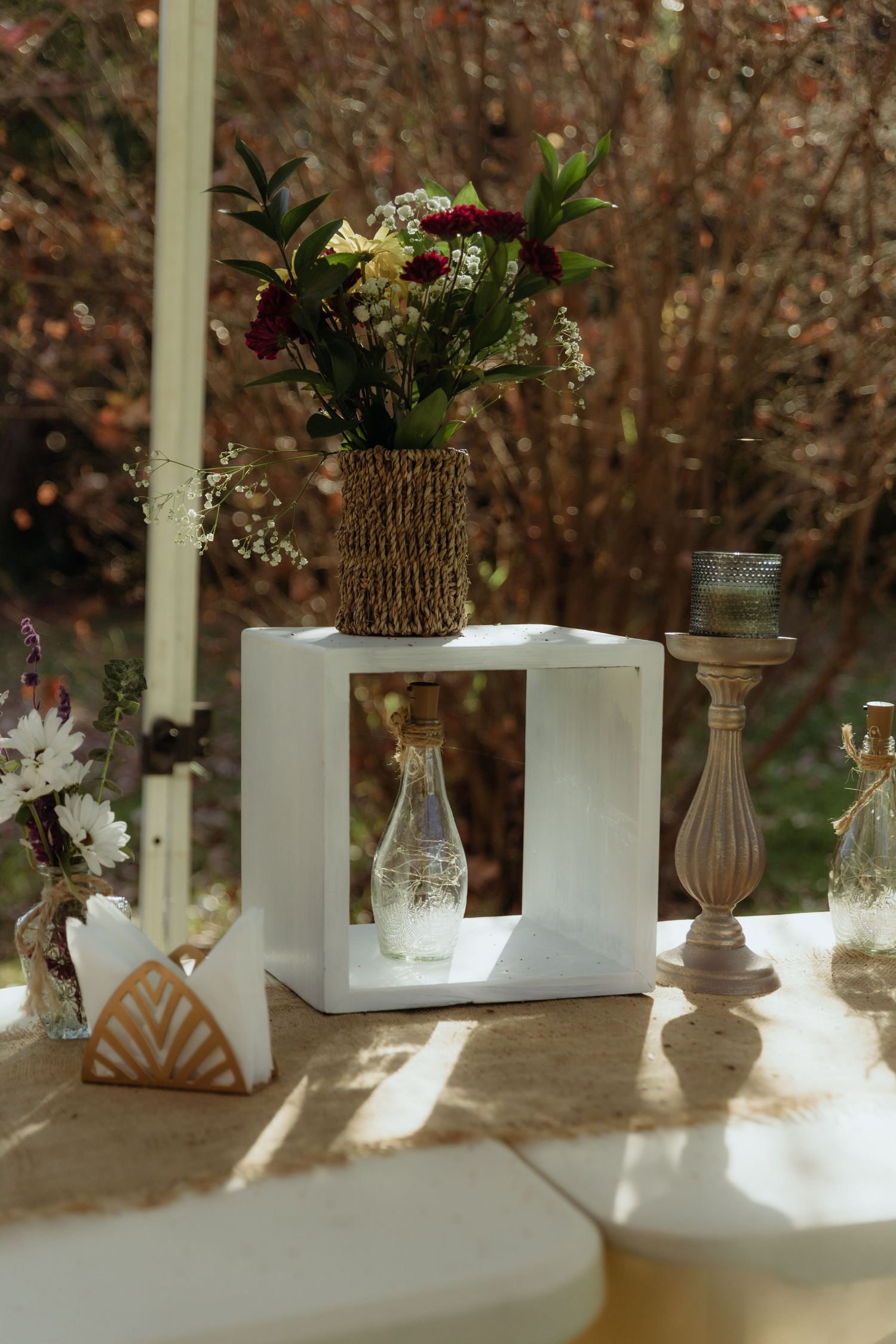 Boho wedding table decor featuring a woven vase with flowers, white cube stand, and glass bottles.