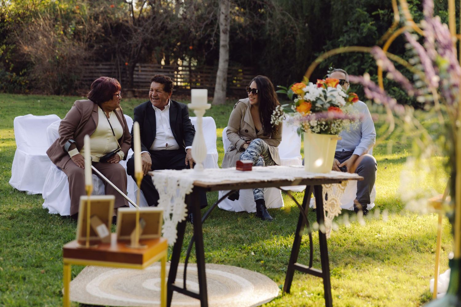 Wedding guests seated outdoors on white chairs near a floral arch during an outdoor garden ceremony.