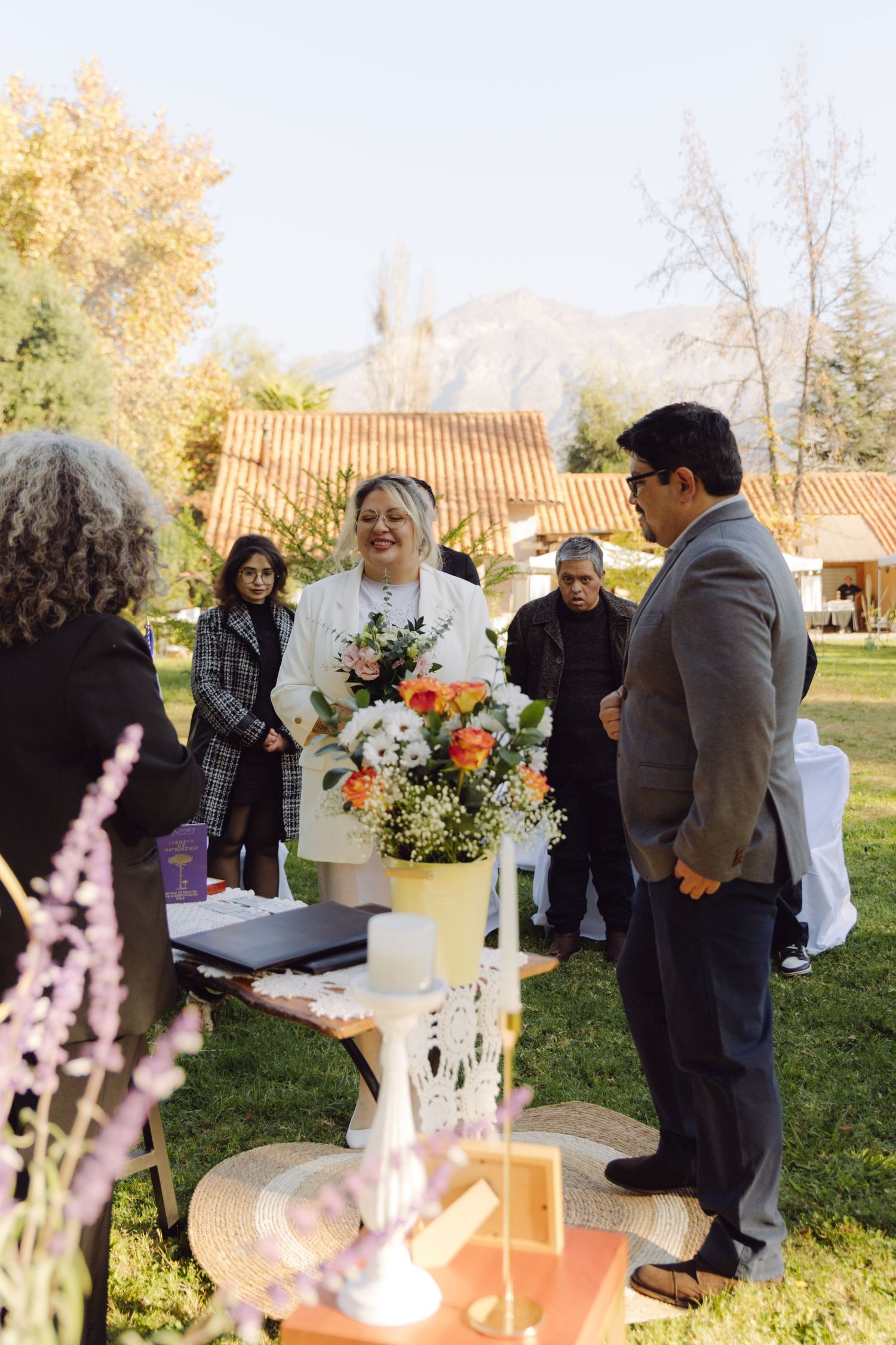 Smiling bride in white blazer holds floral bouquet during outdoor wedding ceremony with guests and mountain backdrop.
