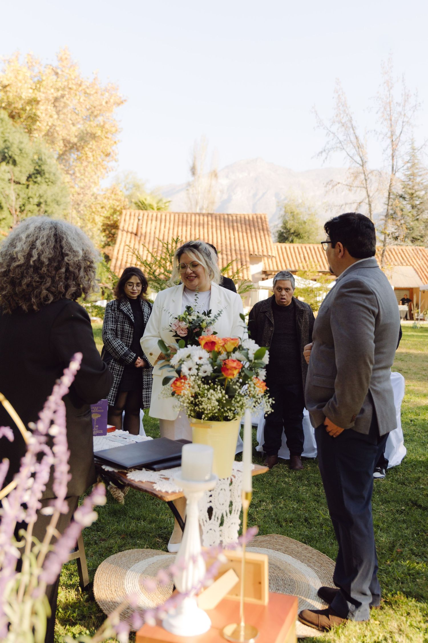 Outdoor wedding ceremony with bride in white suit holding colorful bouquet, groom in gray suit, guests gathered on green lawn.