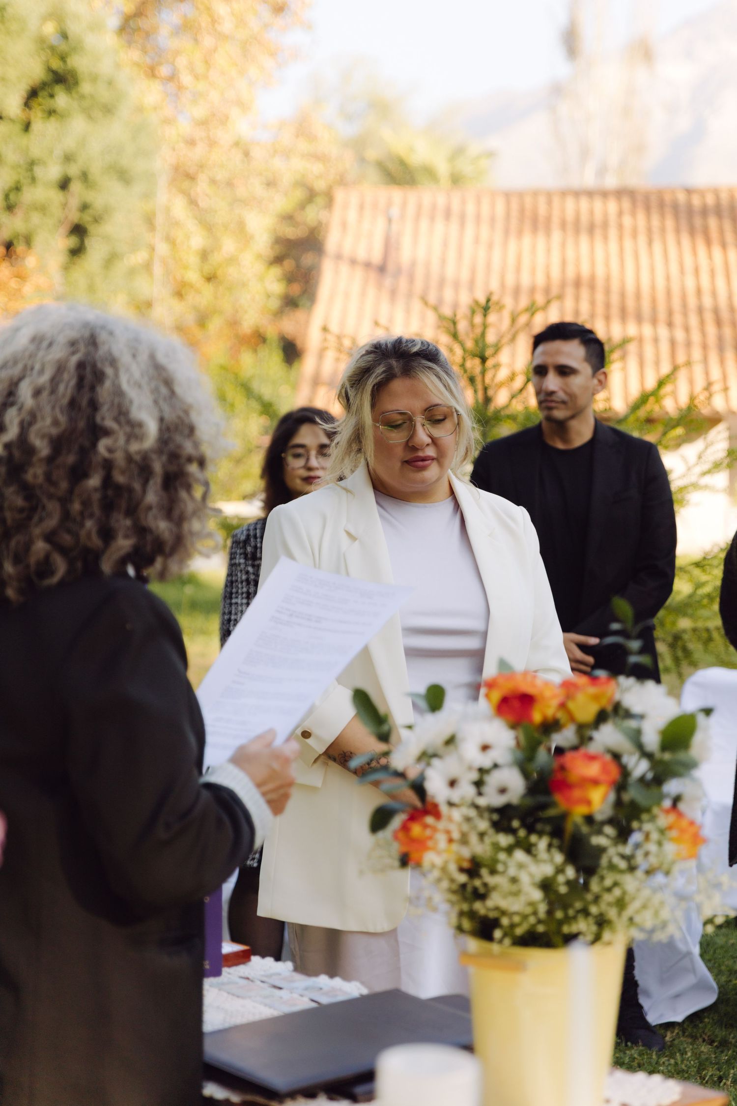 Bride in white blazer receives document during outdoor wedding ceremony surrounded by guests and colorful floral arrangements.