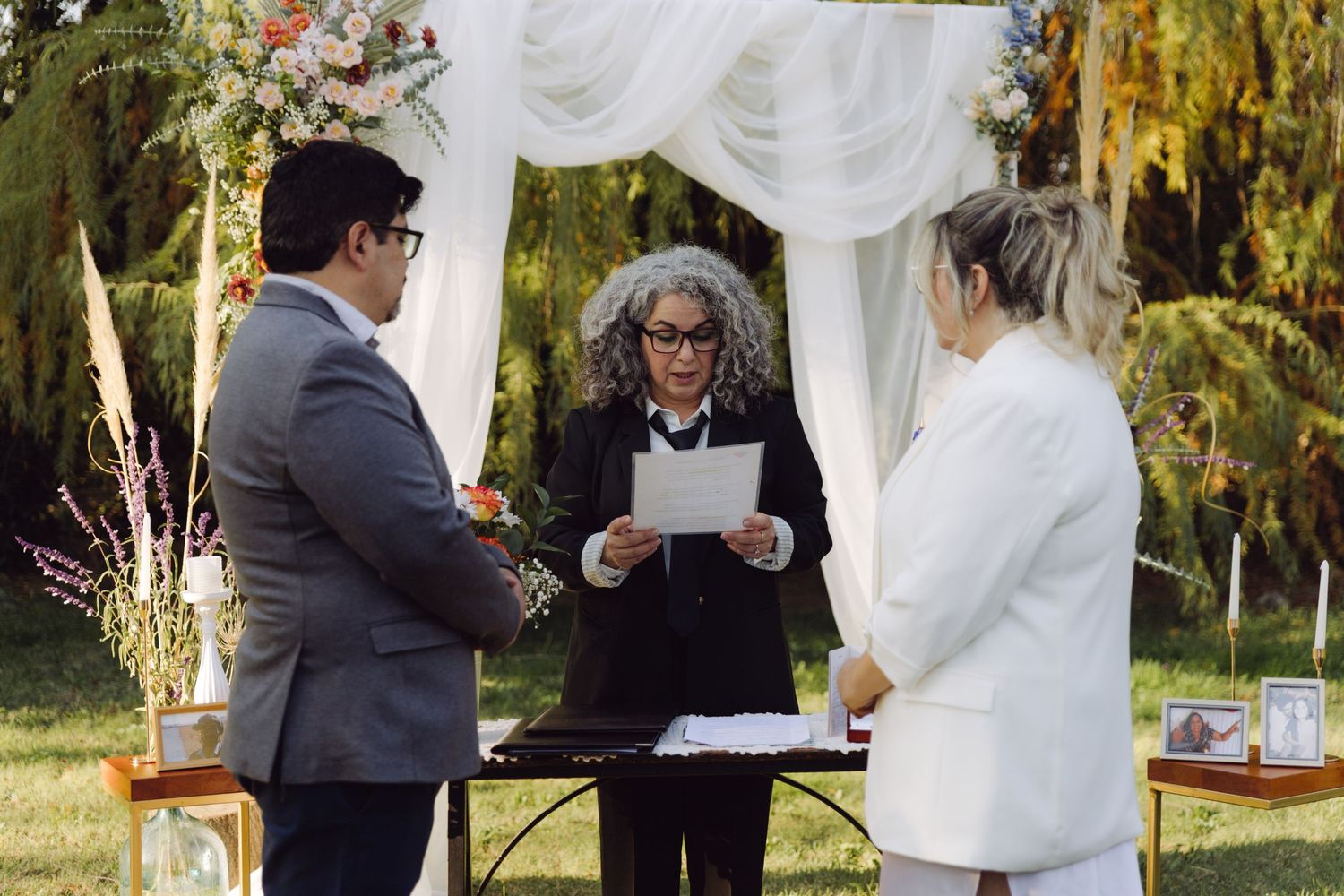 Wedding officiant reads vows to couple standing under white draped arch decorated with flowers in outdoor ceremony.