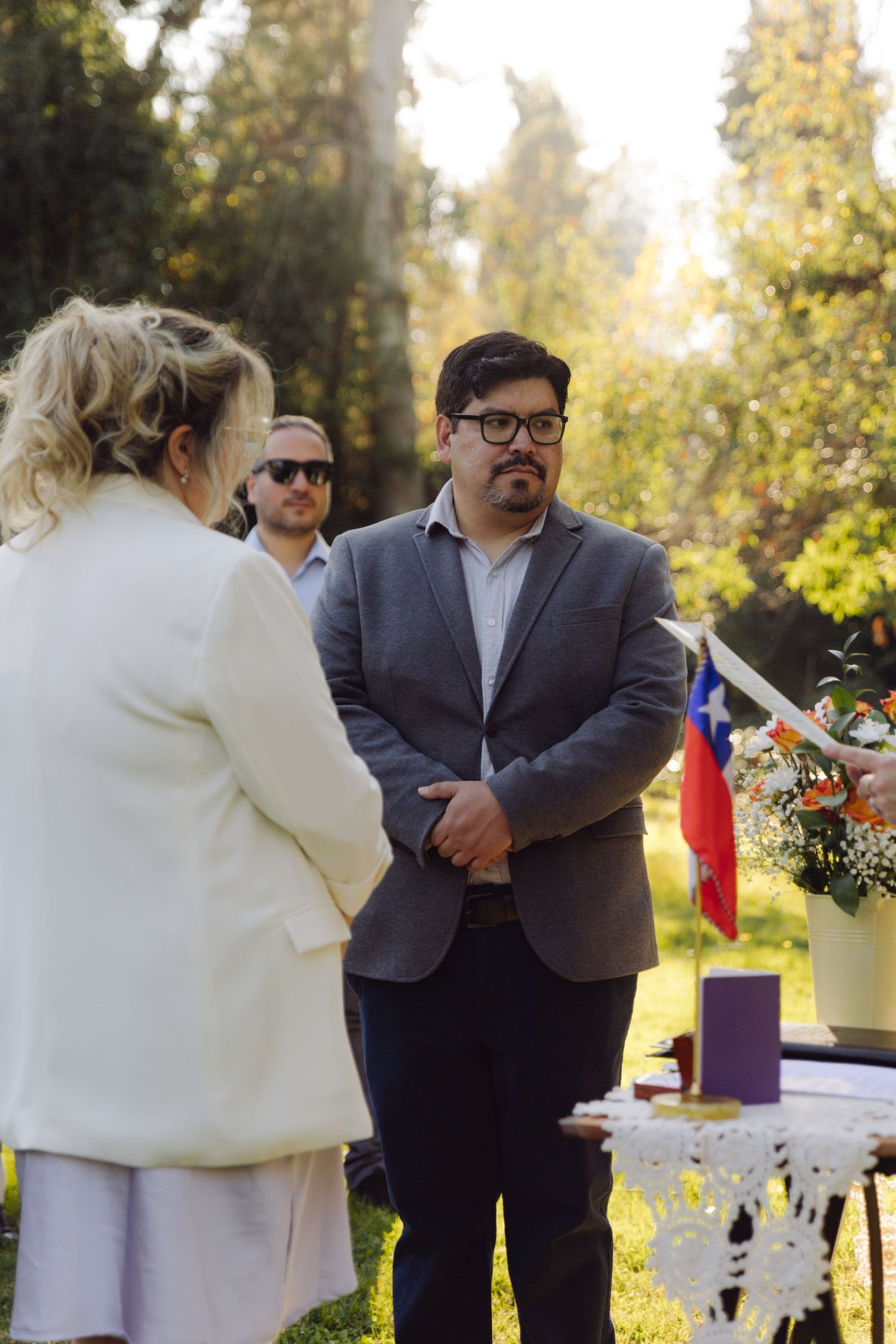 Groom in gray suit listens attentively to bride during outdoor wedding ceremony with Chilean flag visible in background.