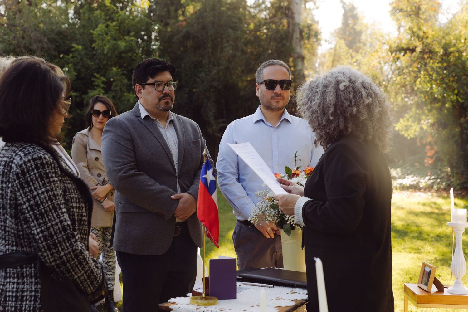 Wedding guests and couple stand near Chilean flag during outdoor ceremony in a sunlit garden setting.