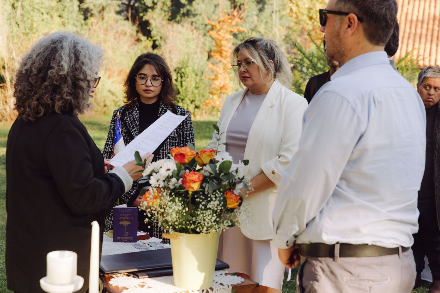 Wedding officiant reads to couple and guests during outdoor ceremony beside orange and white floral centerpiece arrangement.