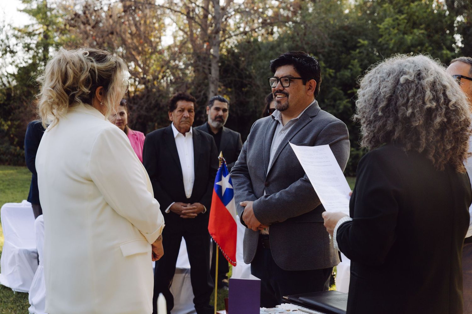 Bride and groom exchange vows outdoors with Chilean flag backdrop, officiant holding papers, guests watching ceremony.
