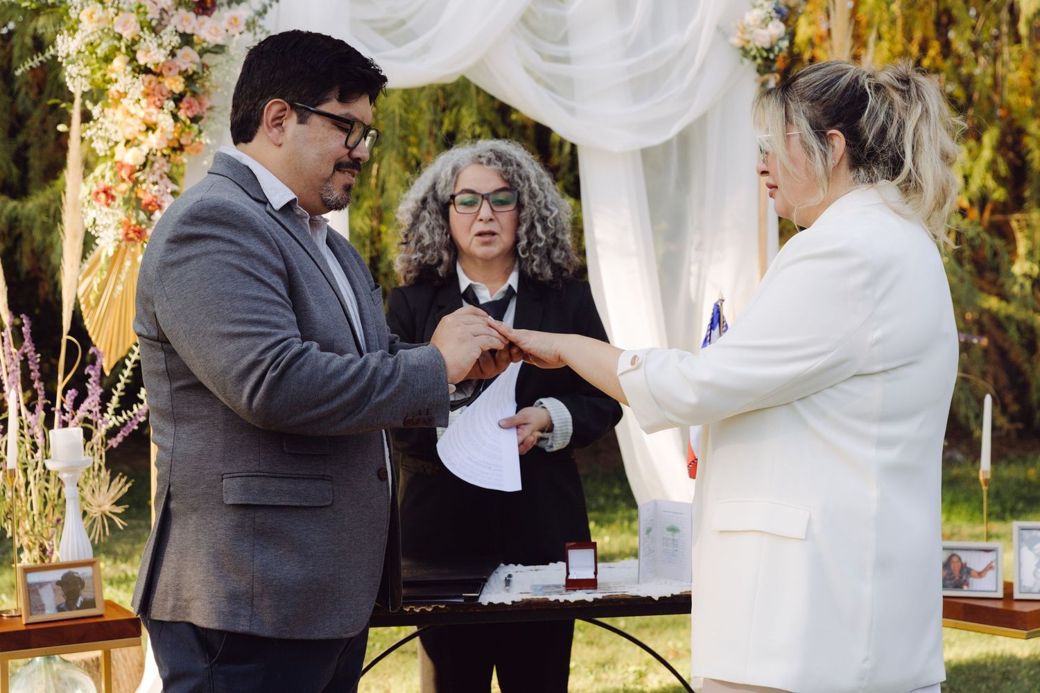 Couple exchanges rings during outdoor wedding ceremony under floral arch with officiant reading from papers behind them.