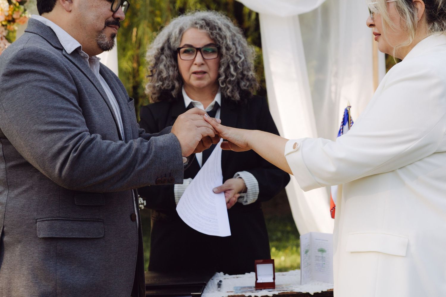 Groom places wedding ring on brides finger as officiant watches during intimate outdoor wedding ring exchange ceremony.