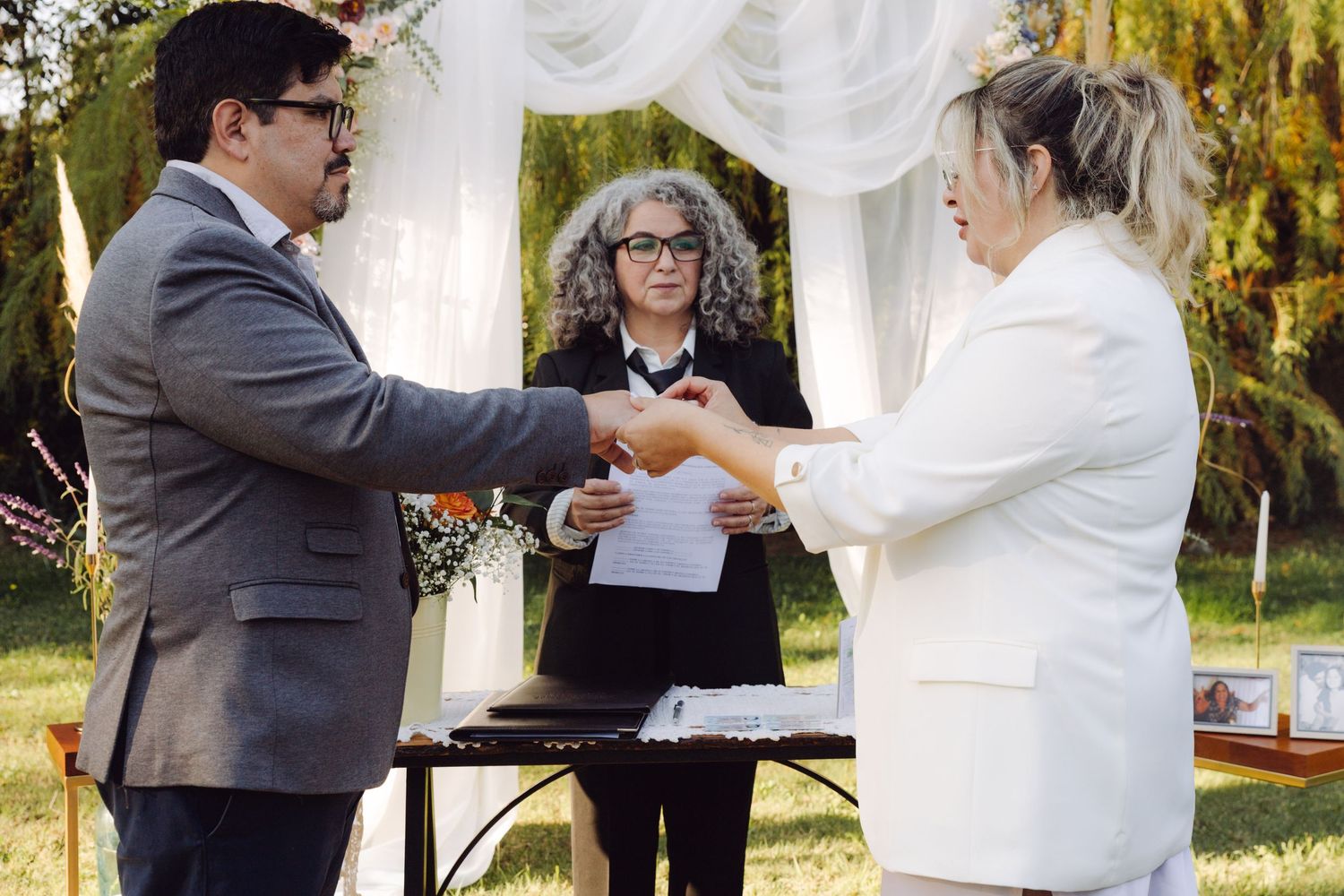 Couple exchanging rings during outdoor wedding ceremony with officiant in garden setting.