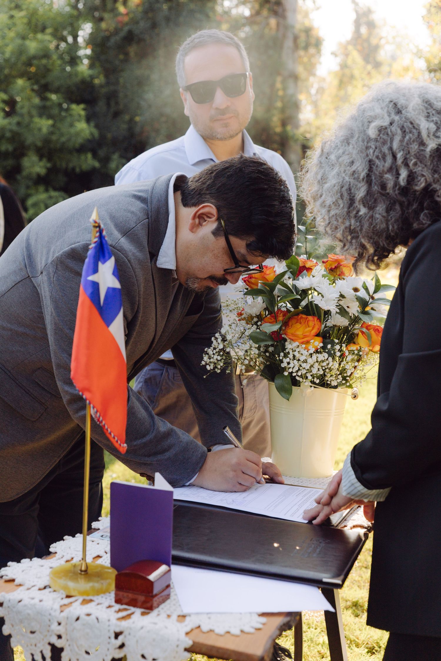 Man signing wedding register at outdoor ceremony table with Chilean flag and flowers.