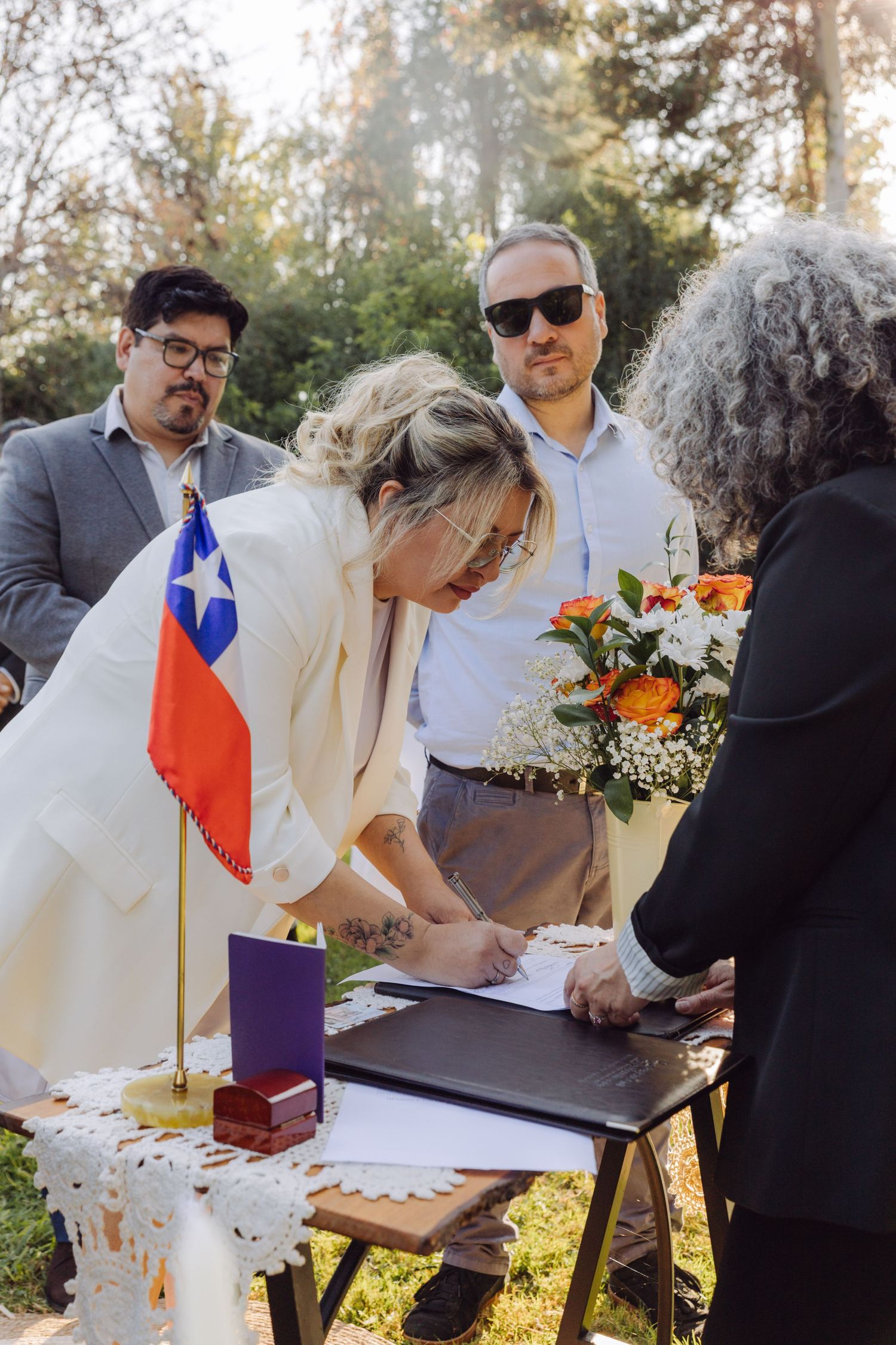 Bride signing marriage documents at outdoor ceremony table with Chilean flag displayed.