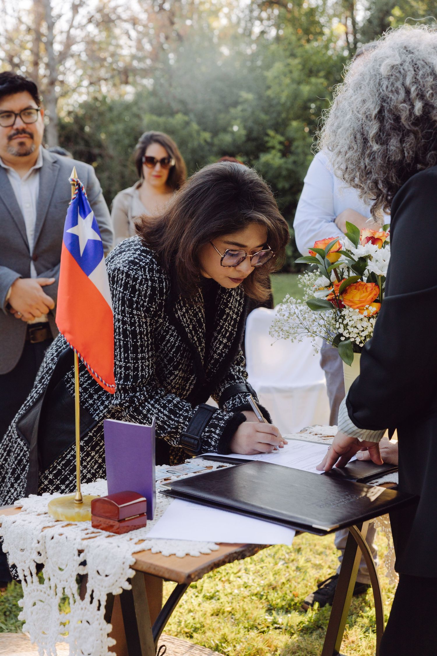 Woman signing wedding register at outdoor Chilean ceremony with flowers and guests nearby.
