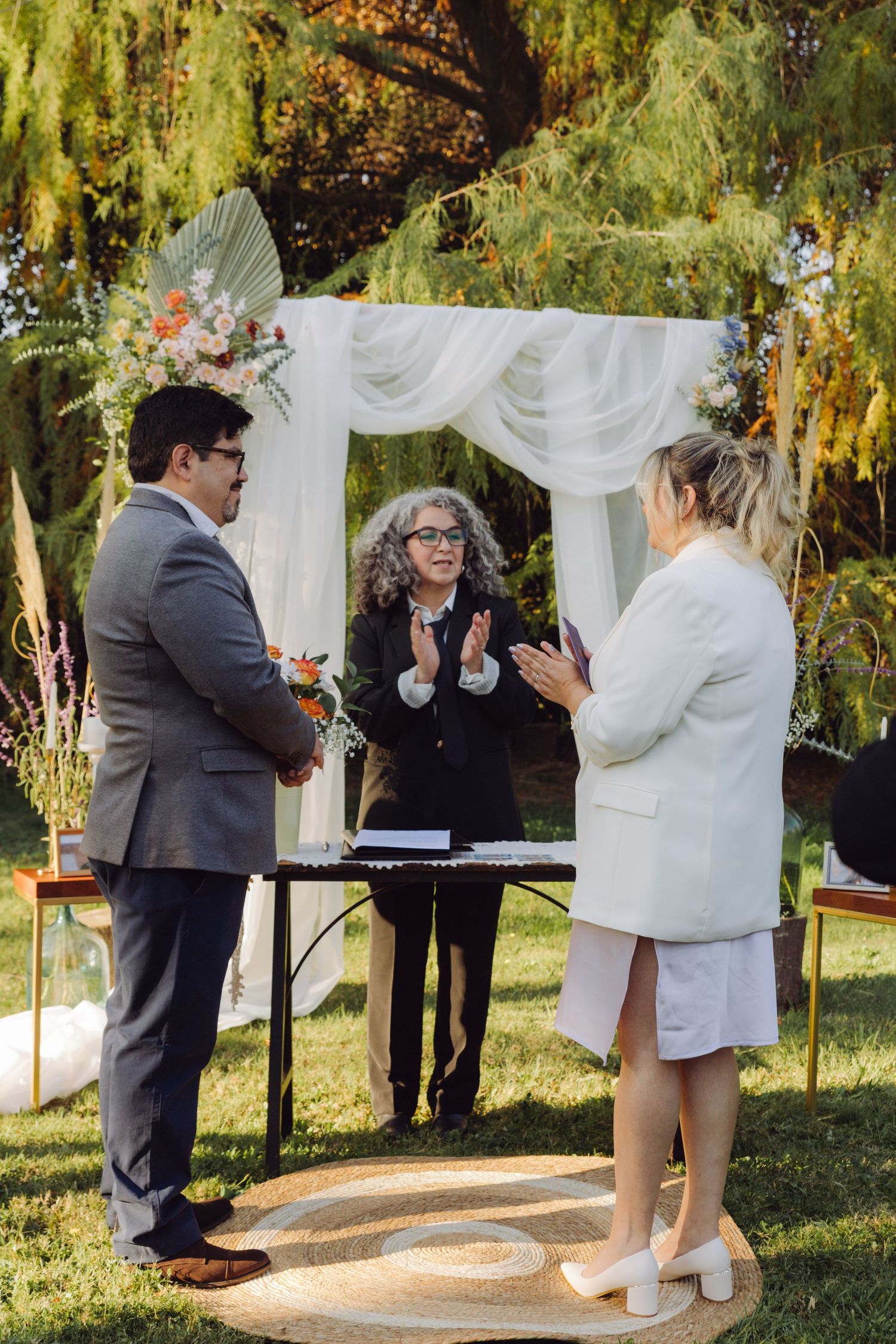 Officiant clapping joyfully as bride and groom celebrate during outdoor wedding ceremony.