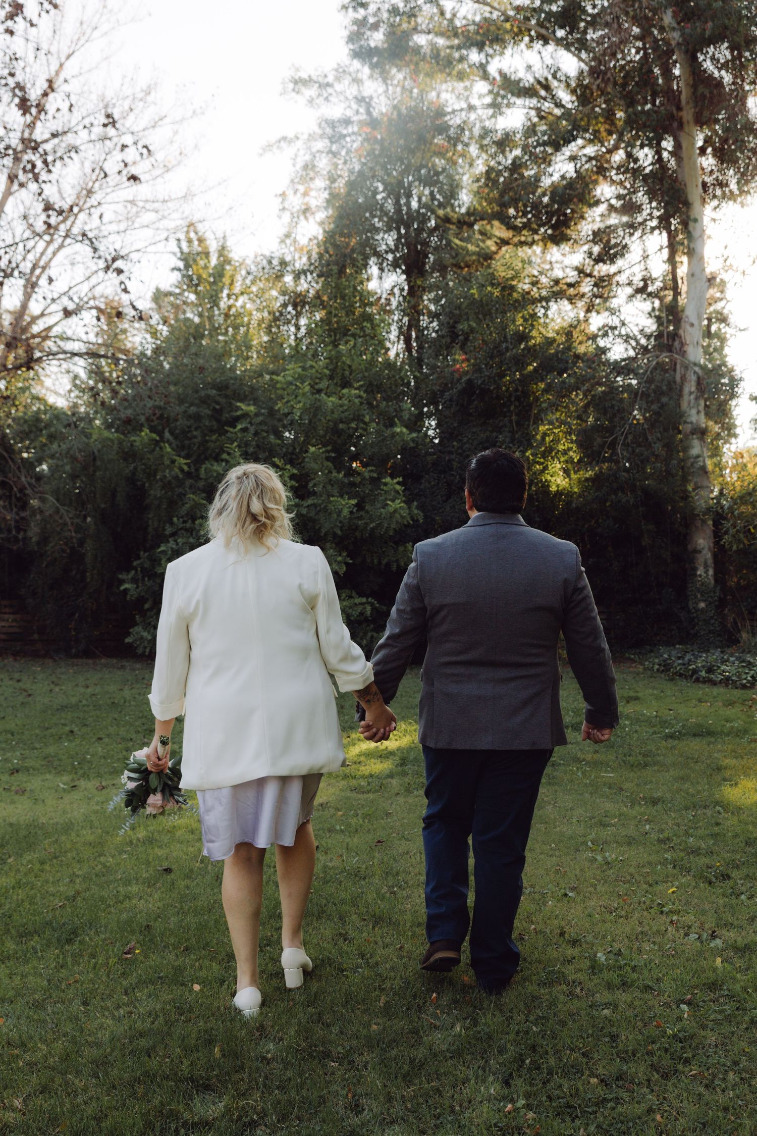 Newlywed couple walks hand in hand across green lawn surrounded by trees.