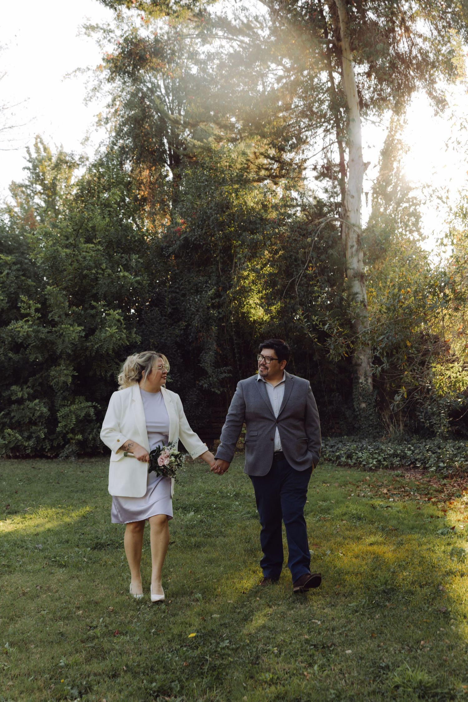 Bride and groom hold hands walking through sunlit garden on wedding day.