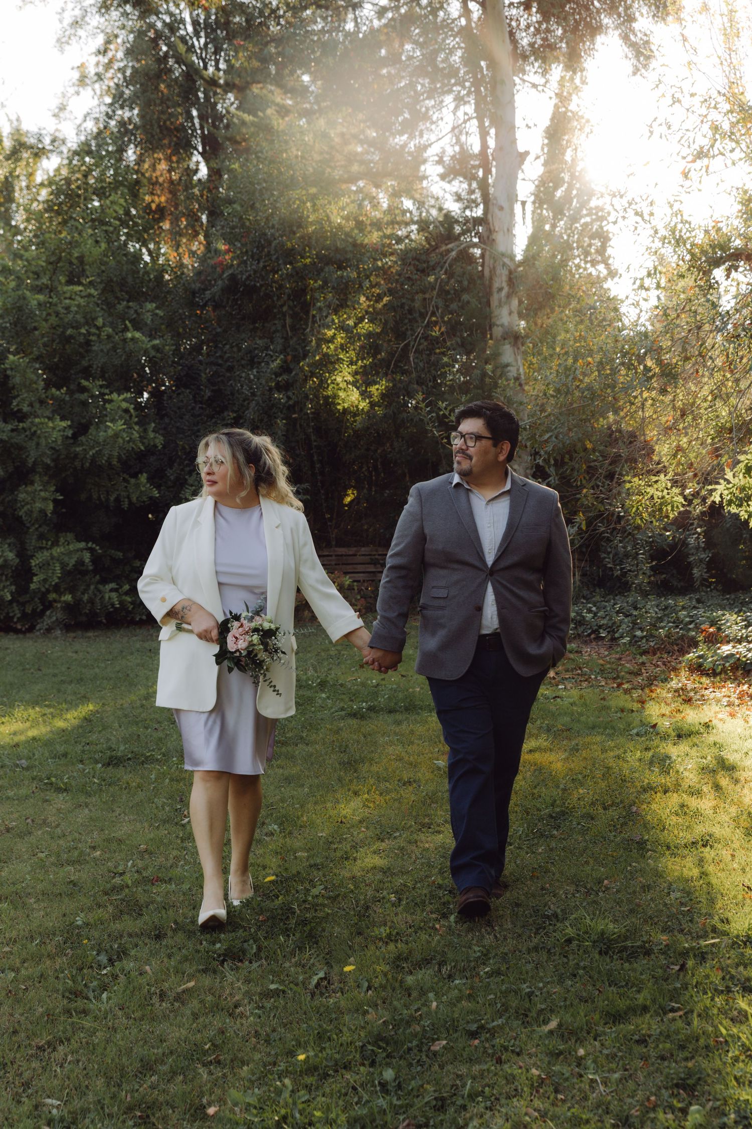 Couple in wedding attire strolls hand in hand through lush garden at sunset.