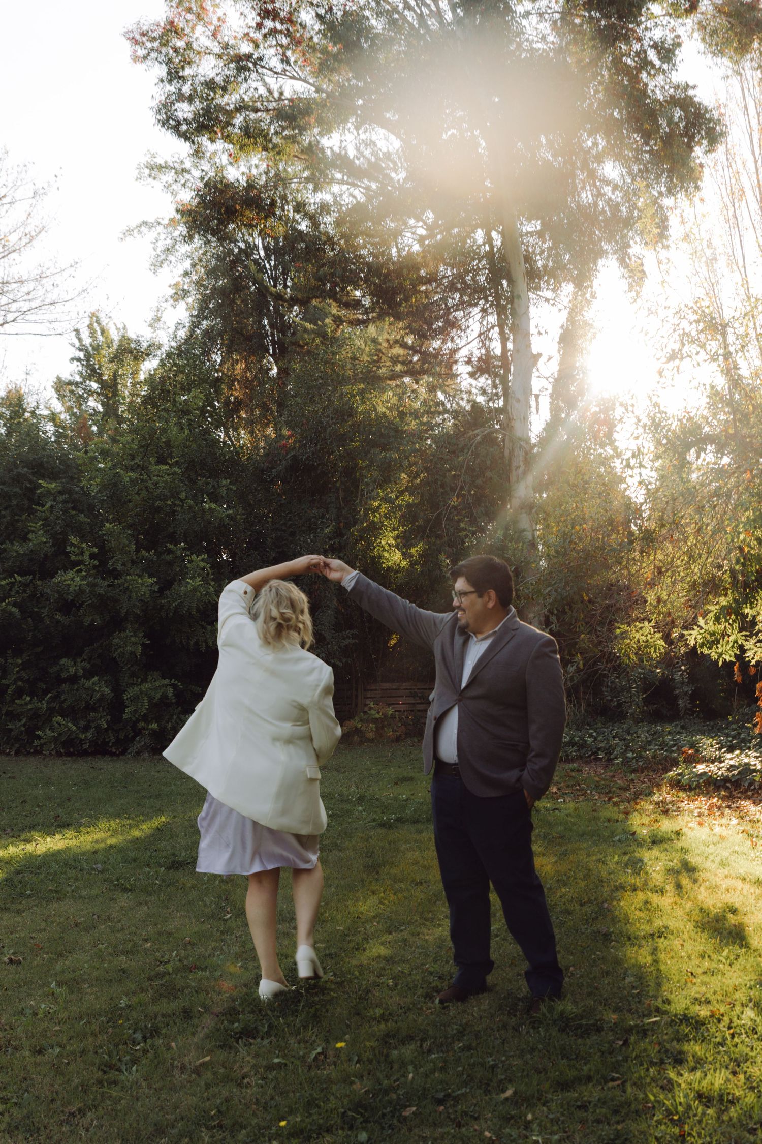 Groom spins bride in white blazer during playful outdoor wedding dance.
