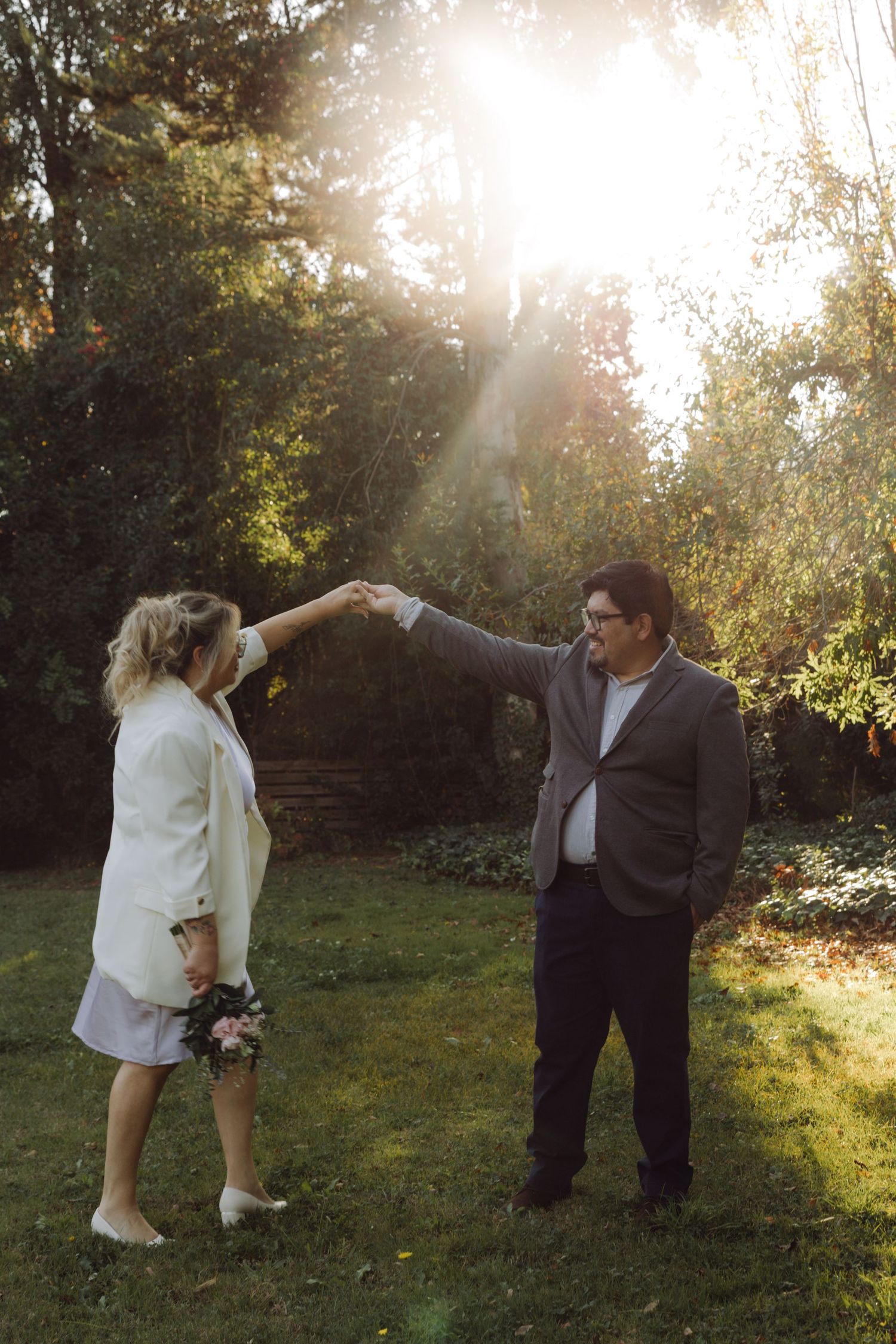 Newlyweds hold hands facing each other in golden sunlit garden setting.
