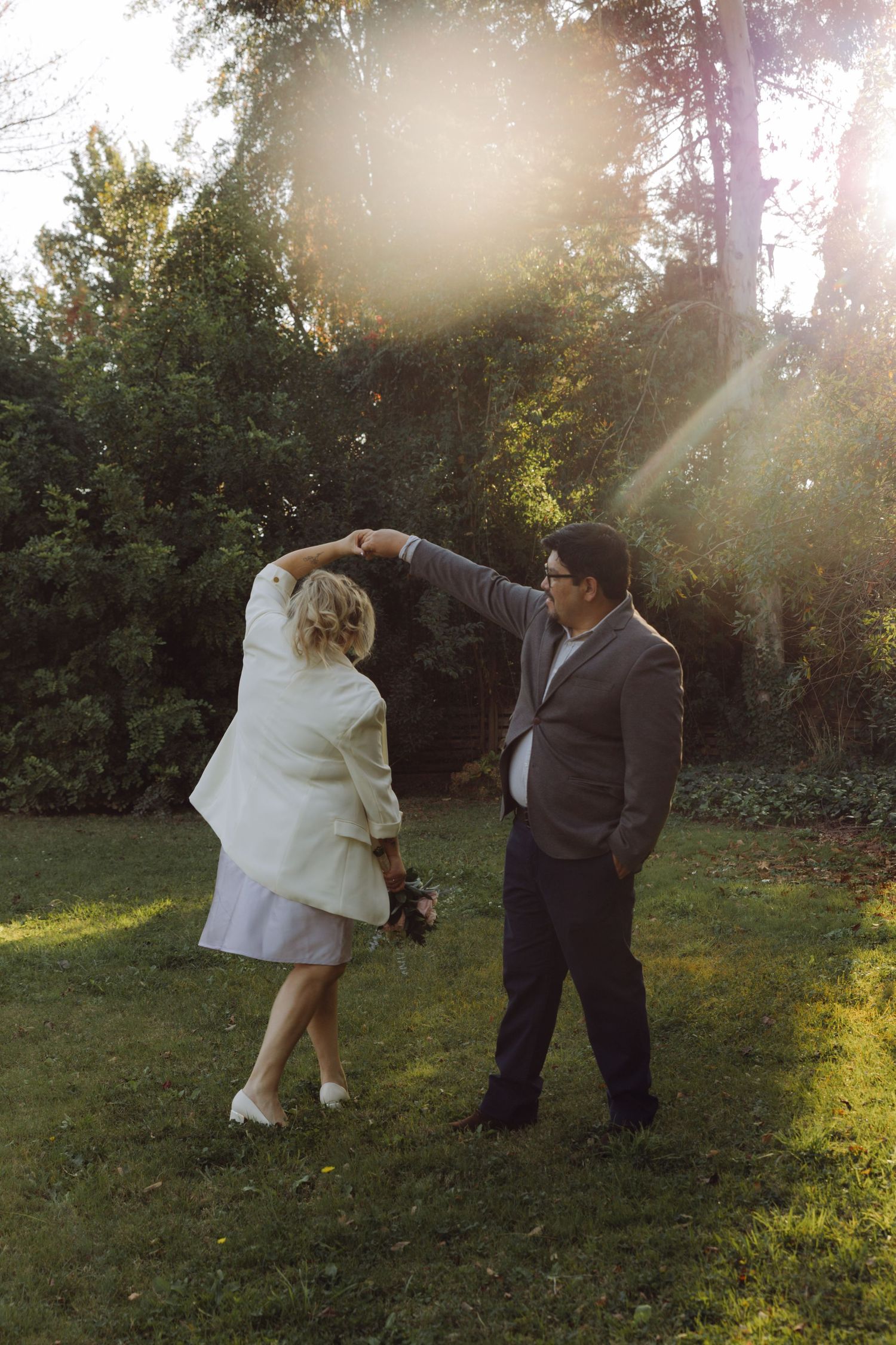 Groom twirls bride holding bouquet in sunlit garden during wedding celebration.