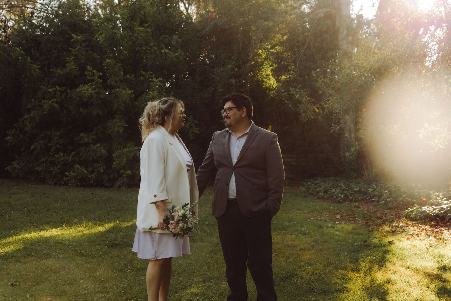 Bride and groom share a smile standing together in glowing sunset garden.