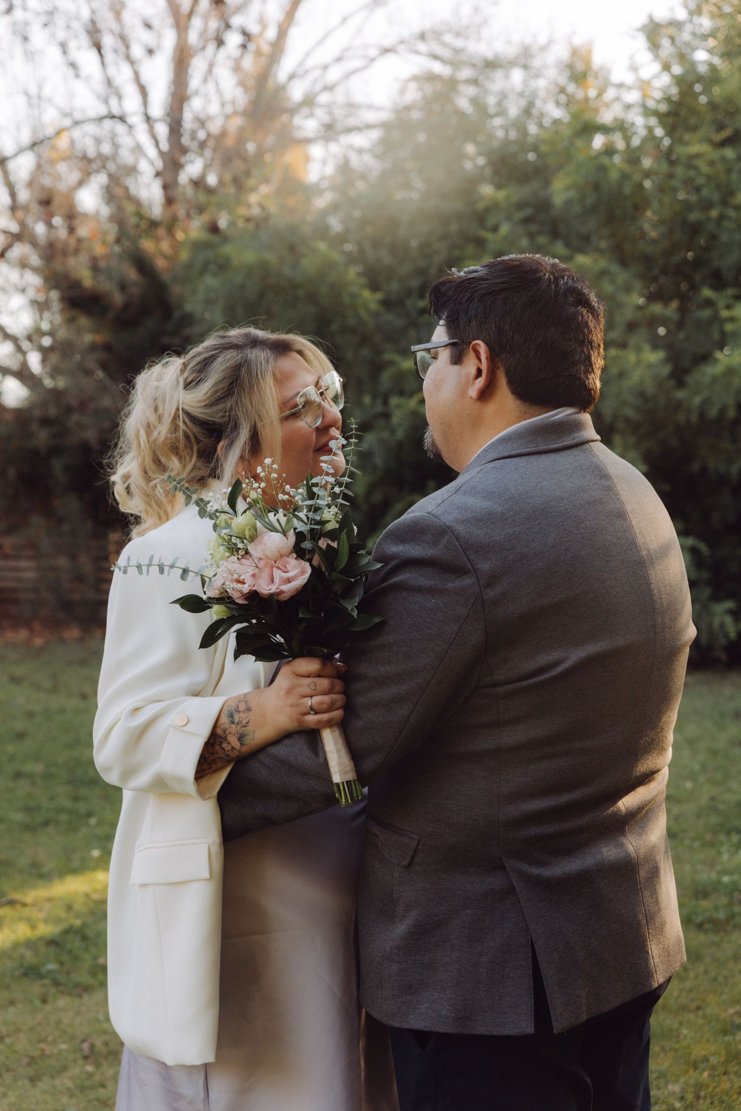 Bride and groom embrace holding pink floral bouquet in outdoor garden setting.