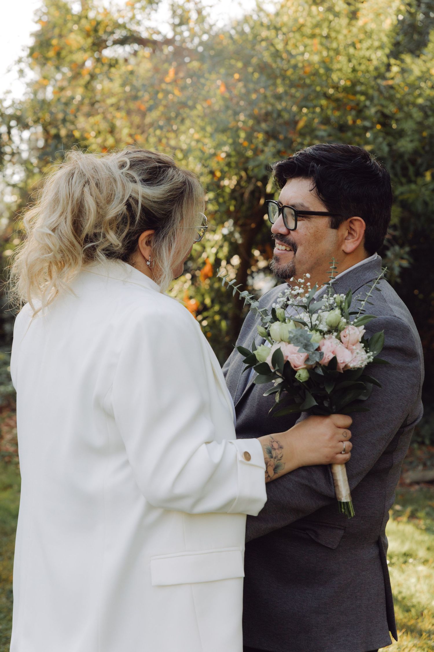 Smiling groom holds bouquet while embracing bride in white blazer outdoors.