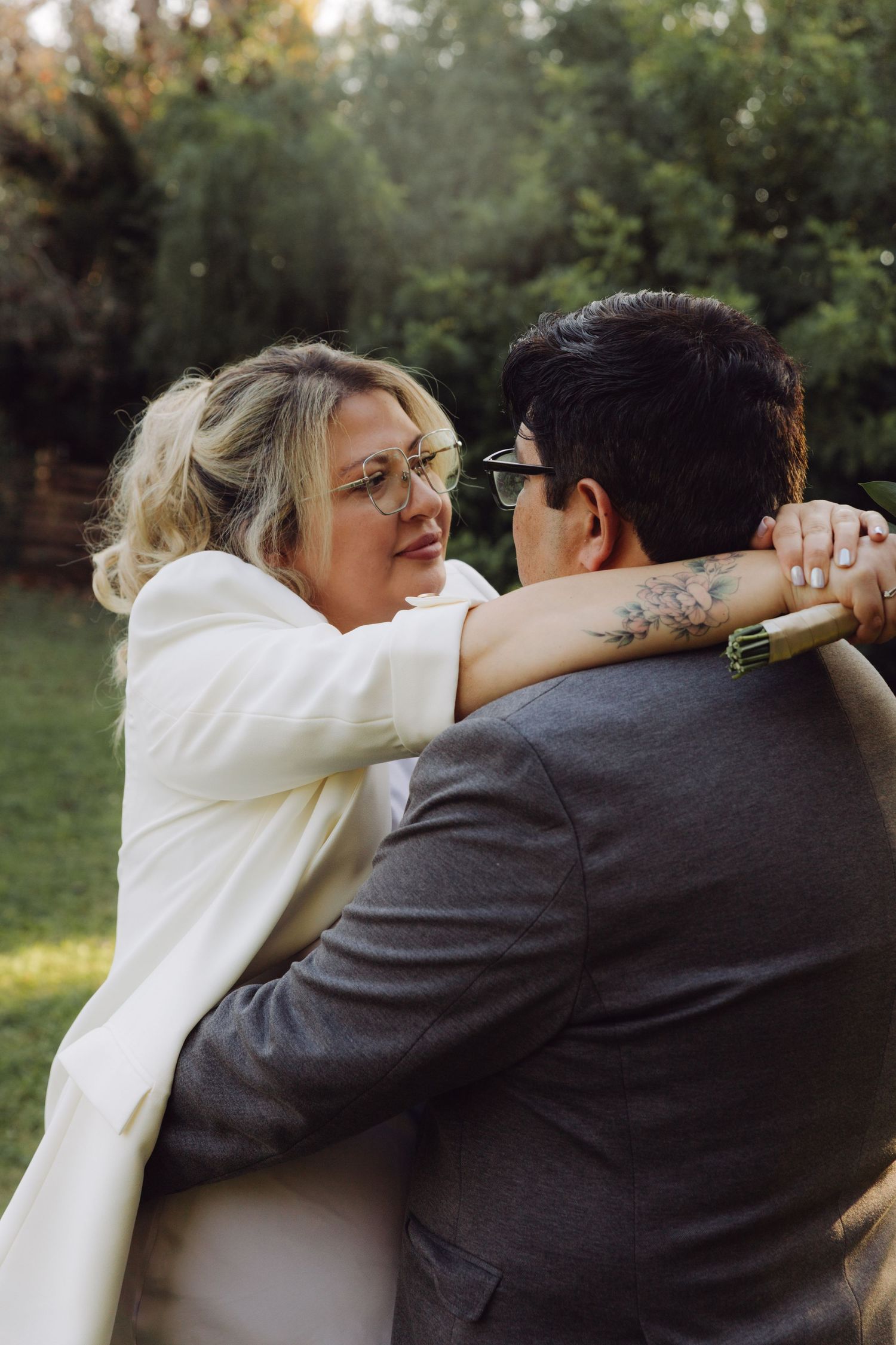 Couple embracing outdoors, woman in white jacket with tattoos hugging man in gray suit in garden setting.