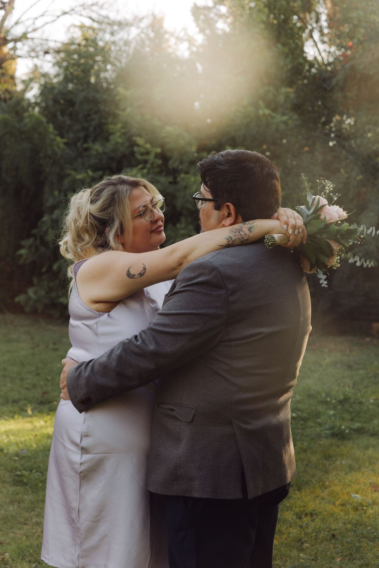 Bride and groom sharing emotional hug outdoors, woman holding pink bouquet with sun flare in background.