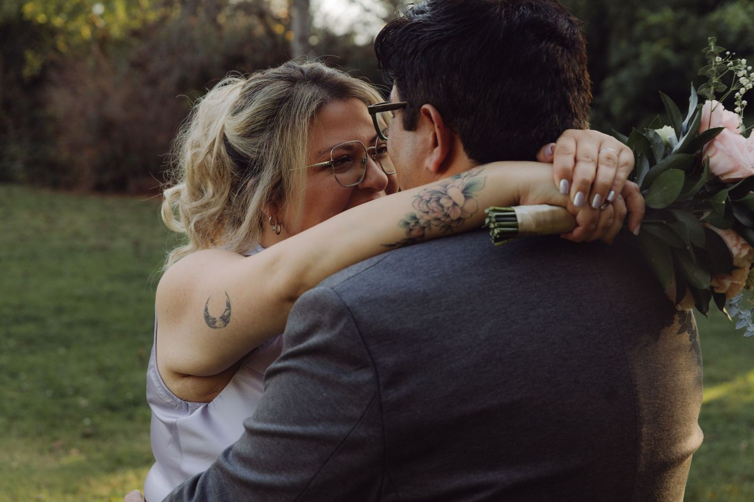 Close-up of couple embracing at wedding, bride with glasses and tattoos hugging groom holding floral bouquet.