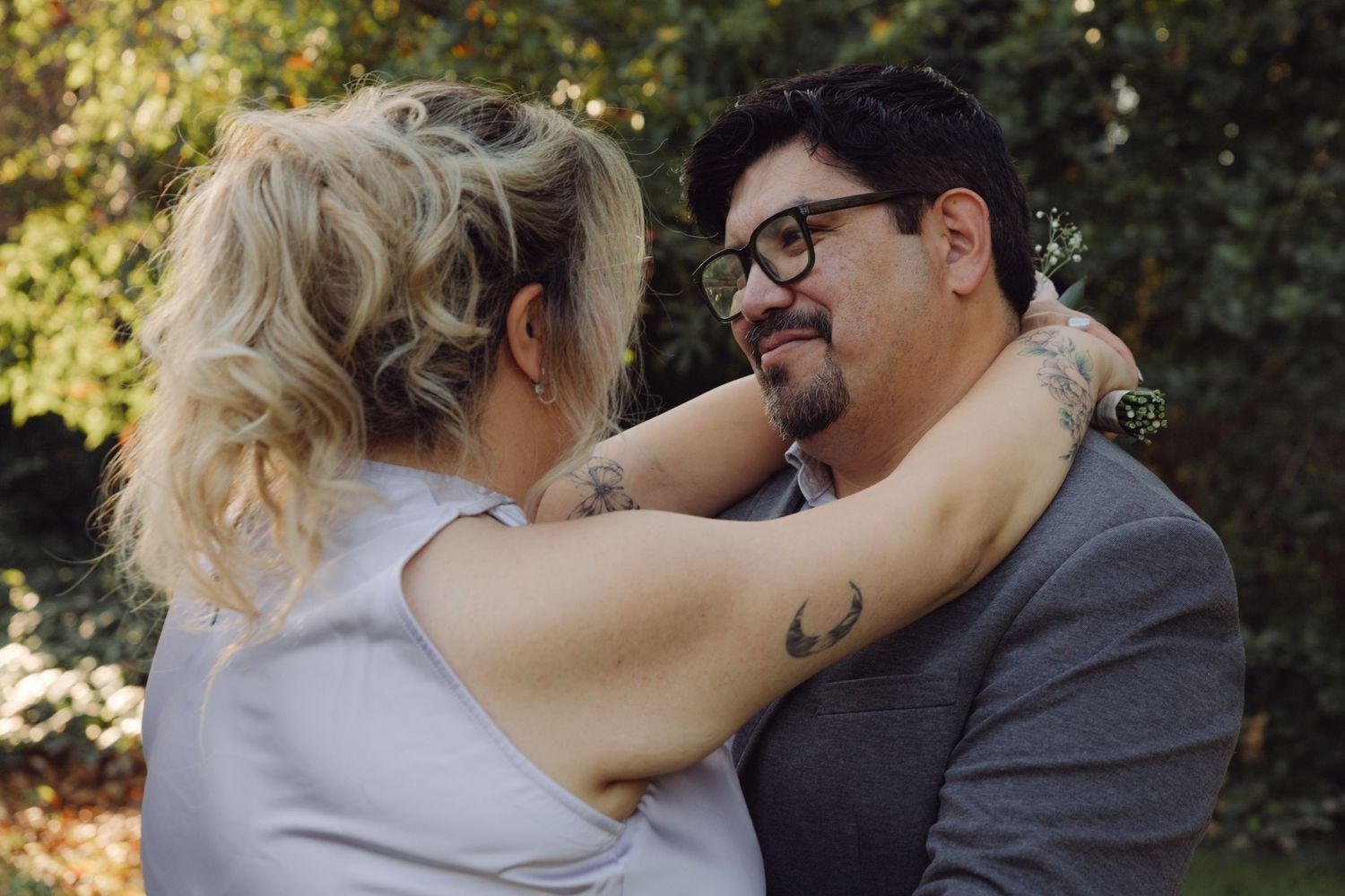 Couple with tattoos sharing intimate moment outdoors, woman embracing man in gray suit among autumn trees.