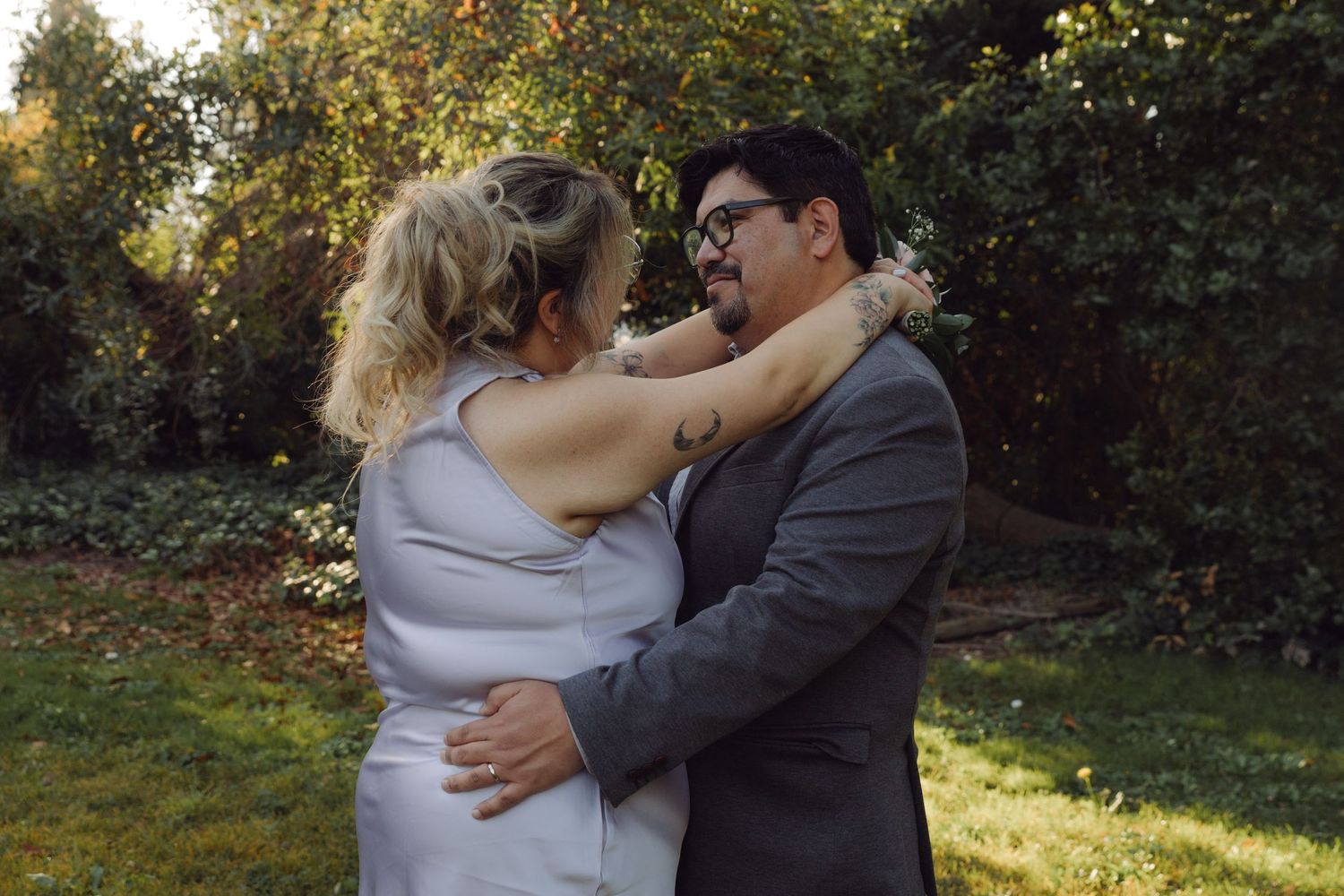Wedding couple embracing in garden, bride in silver dress and groom in gray suit surrounded by fall foliage.
