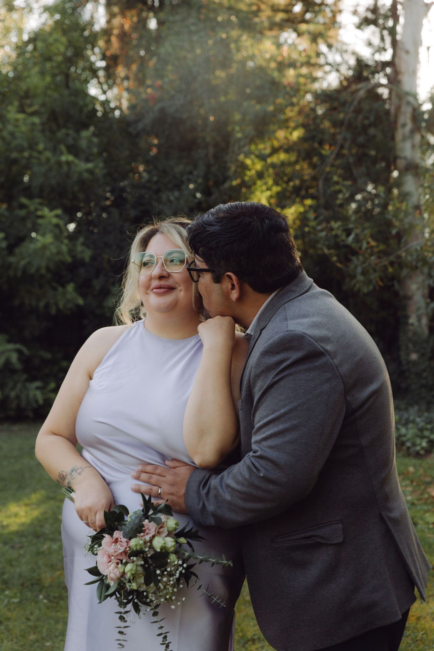 Groom tenderly kisses bride wearing glasses and lavender dress holding pink bouquet in garden setting.