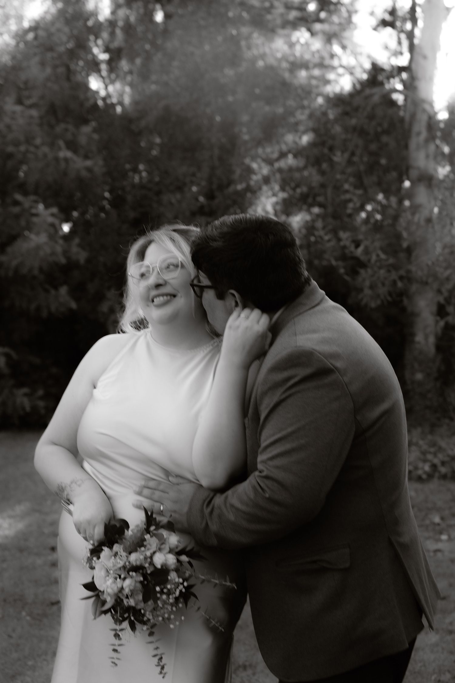 Black and white photo of a groom kissing his smiling bride on the cheek outdoors.