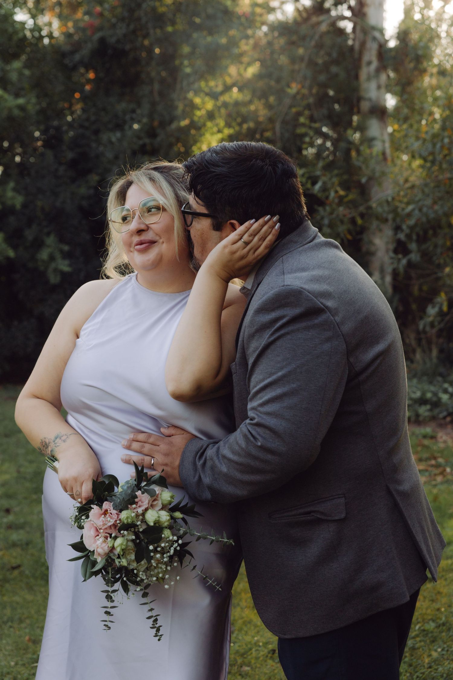 Groom in gray suit kissing bride on cheek as she smiles holding a pink floral bouquet.