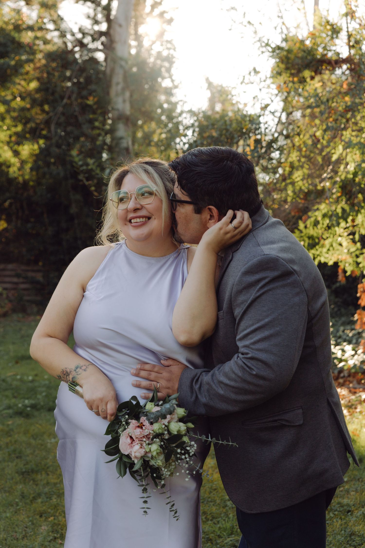 Couple sharing a tender moment outdoors, groom kissing bride's cheek as she laughs in sunlight.